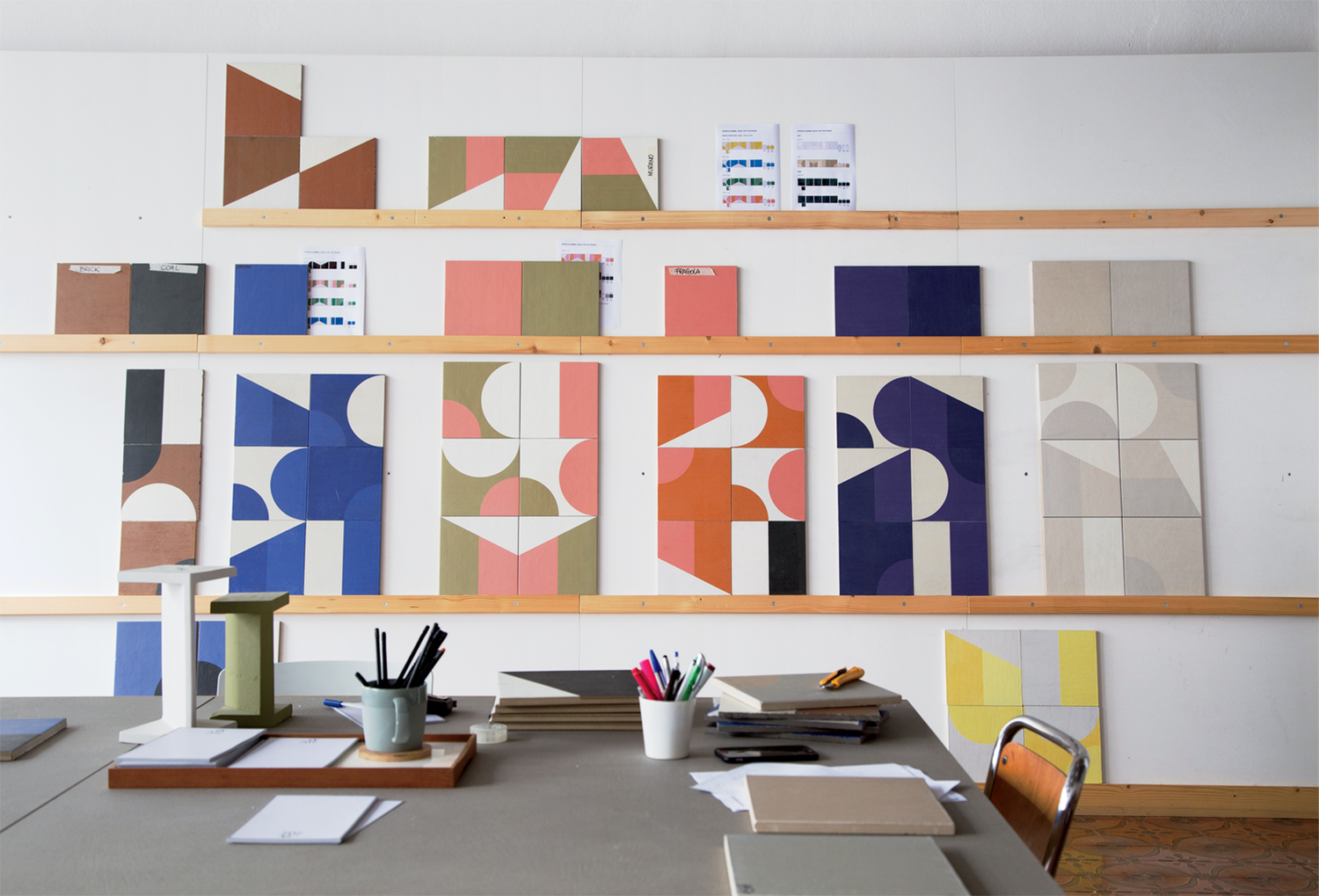 A studio workspace with geometric patterns and colored samples displayed on shelves above a desk scattered with office supplies and papers.