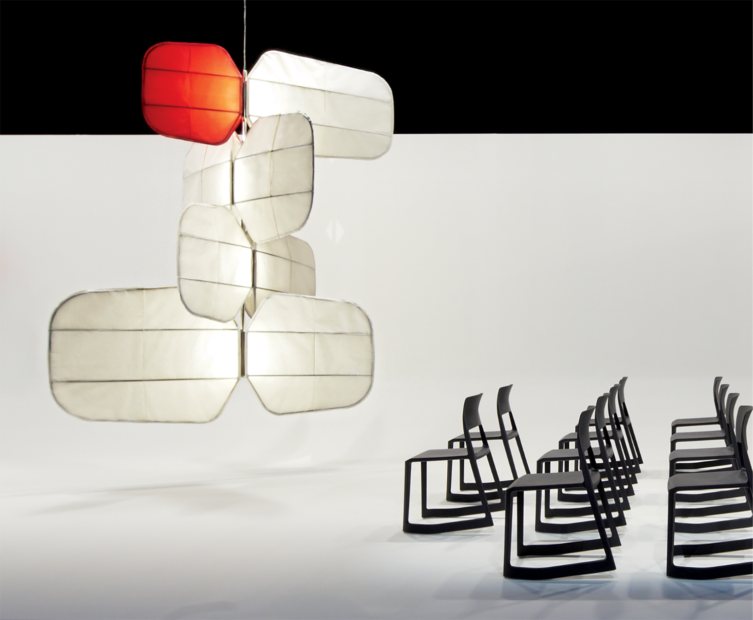 A group of modern black chairs faces large, abstract white and red hanging light fixtures in a minimalist white room with a black upper background.