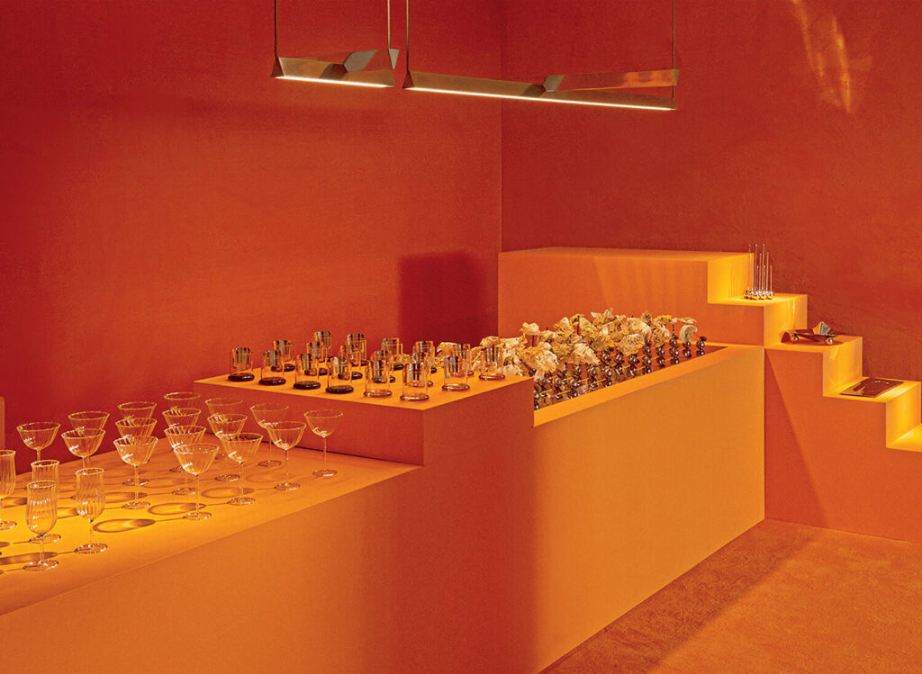 A room with orange walls and yellow platforms displaying glassware, metallic objects, and decorative items, with modern ceiling lights above.