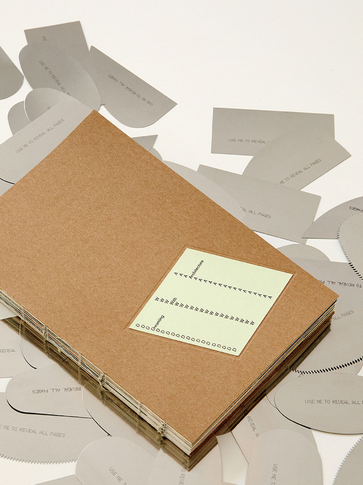 A brown-covered book with a green label lies on a surface surrounded by scattered gray paper tabs labeled “USE ME TO REVEAL ALL PAGES.”.