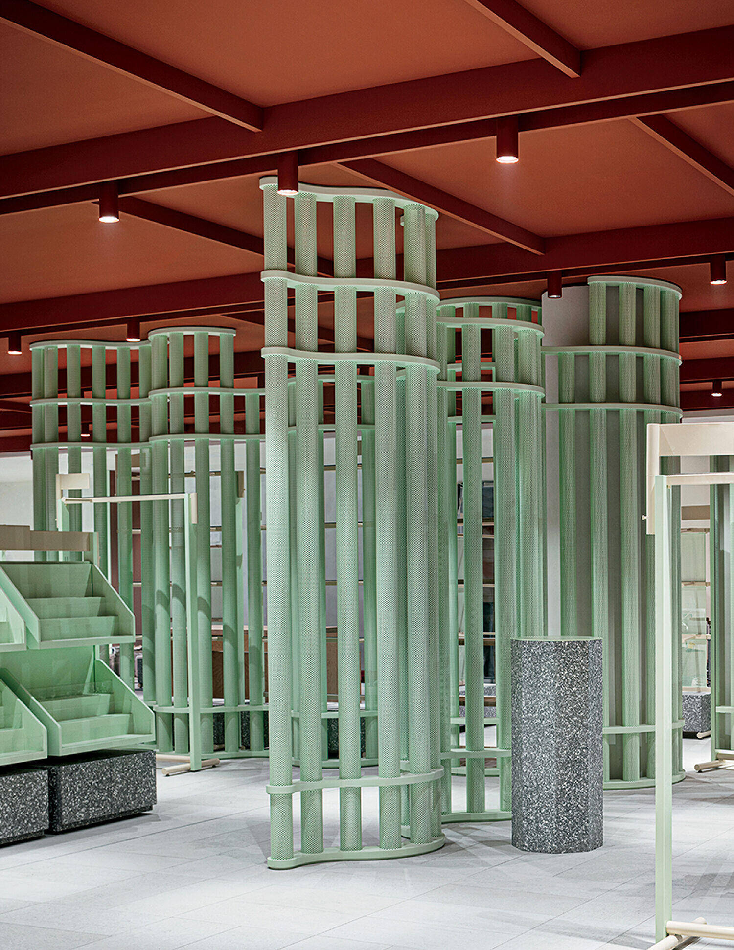 Green cylindrical metal structures stand on a grey tiled floor under a red ceiling with exposed beams and recessed lights. Geometric shelves and granite columns are also visible in the space.