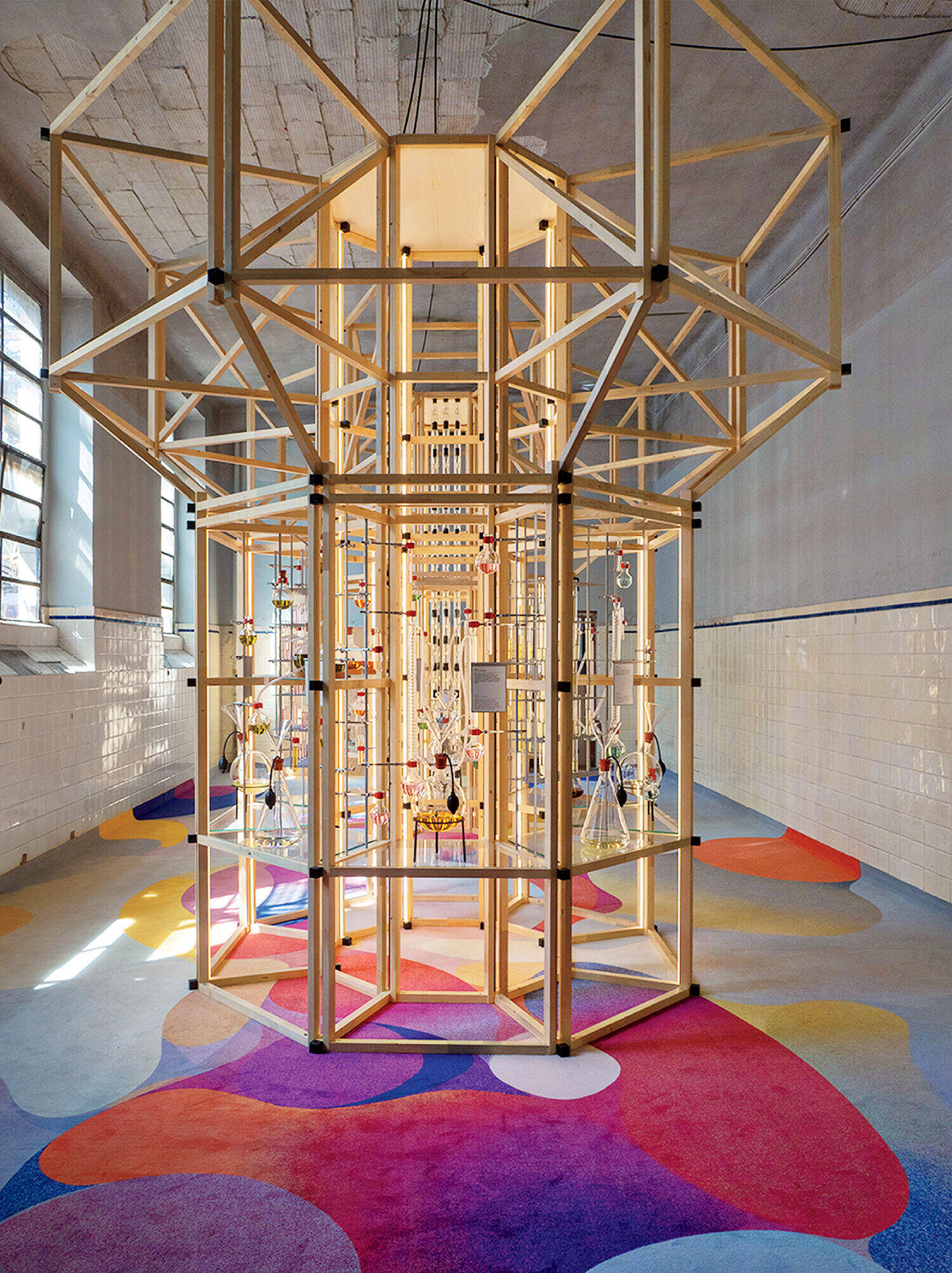Geometric wooden structure with glass tubes and flasks stands in a tiled room with colorful patterned carpet and large windows.