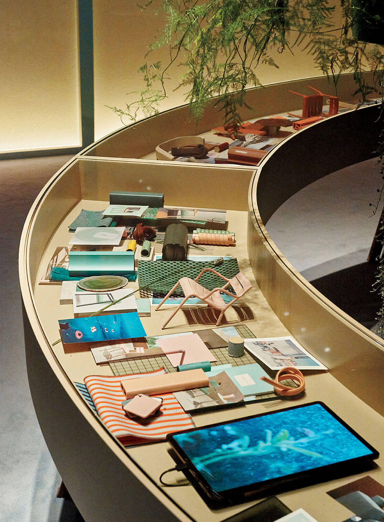 Curved display table with neatly arranged stationery, textiles, objects, and a tablet showing an underwater scene, set in a well-lit room with some greenery.