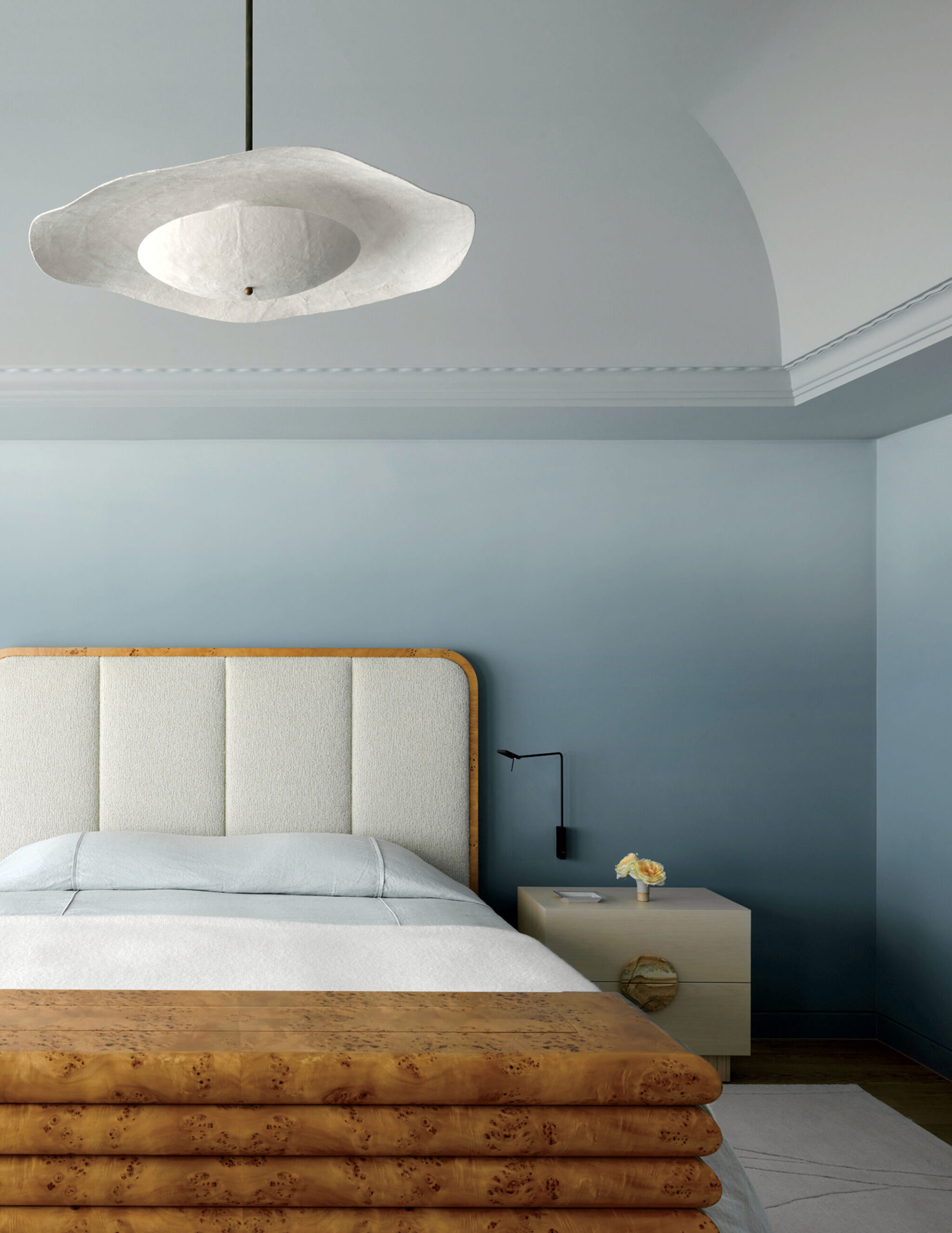 A minimalist bedroom with a padded headboard, wooden footboard, light blue bedding, a white pendant lamp, and a side table with a small flower vase and wall-mounted lamp.