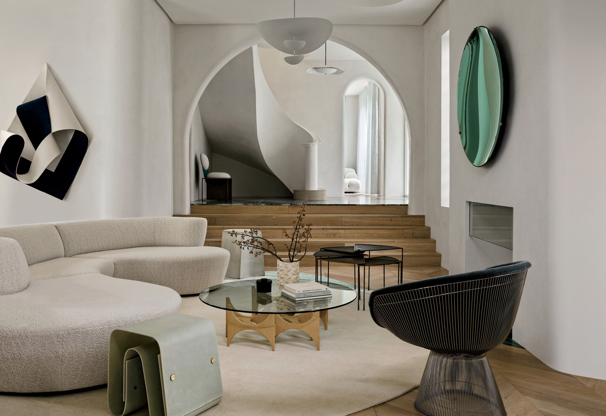 Modern living room with curved beige sofa, glass coffee table, black chair, abstract wall art, large round mirror, arched doorway, and wooden steps leading to a sculptural white staircase.