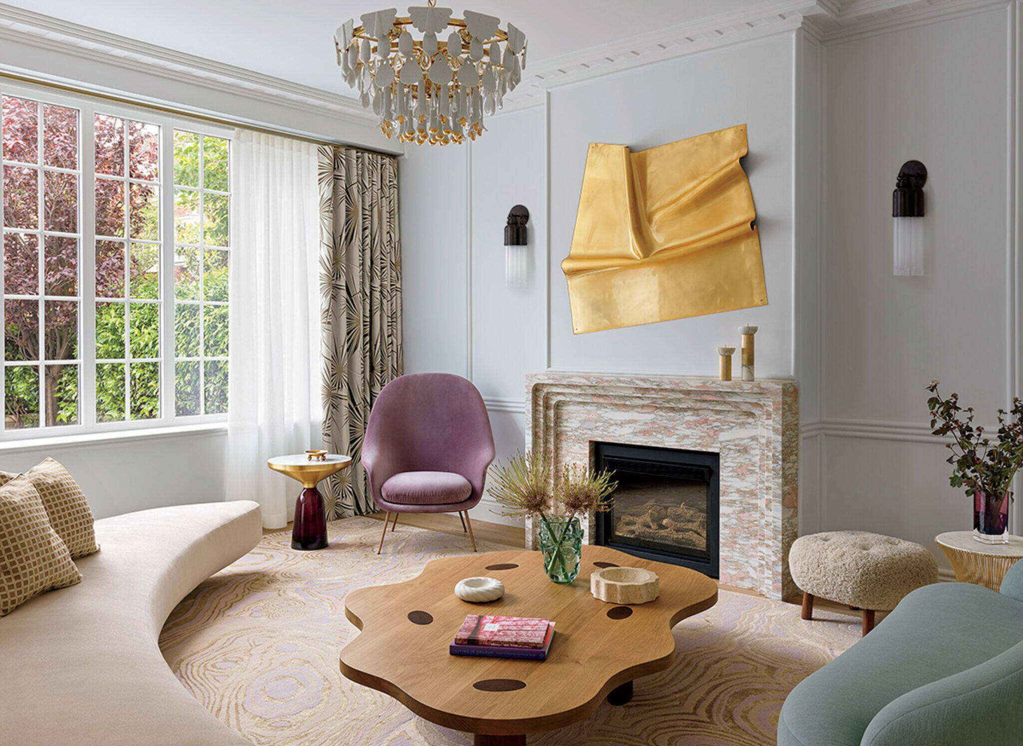 Modern living room with curved sofa, abstract wooden coffee table, purple chair, marble fireplace, gold wall art, and large window with patterned curtains.