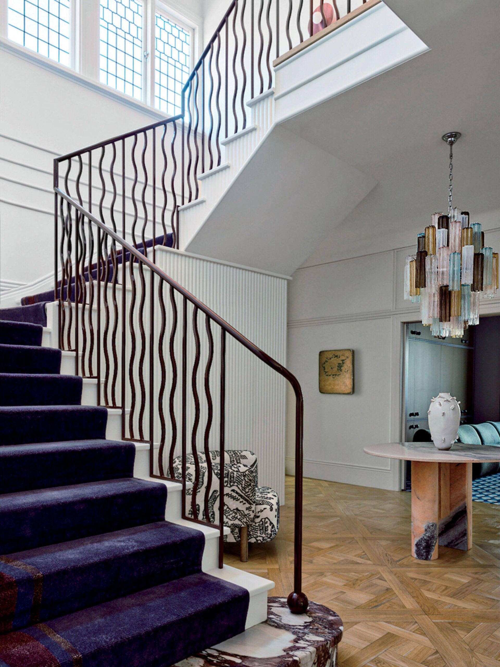 A staircase with a dark wavy railing, navy carpet, and patterned armchair; geometric chandelier hangs over a parquet floor in a modern interior.