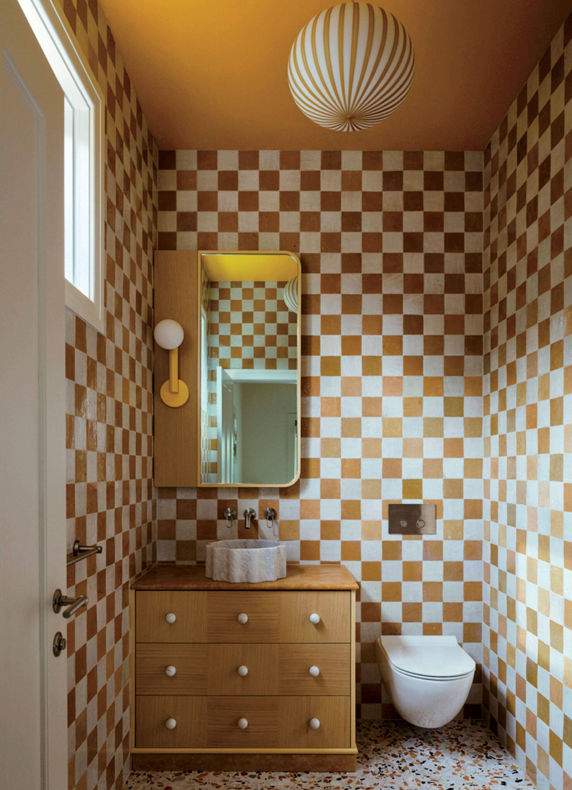 Bathroom with brown and white checkerboard tiles, wooden vanity with marble sink, round light fixture, wall-mounted toilet, and terrazzo floor.
