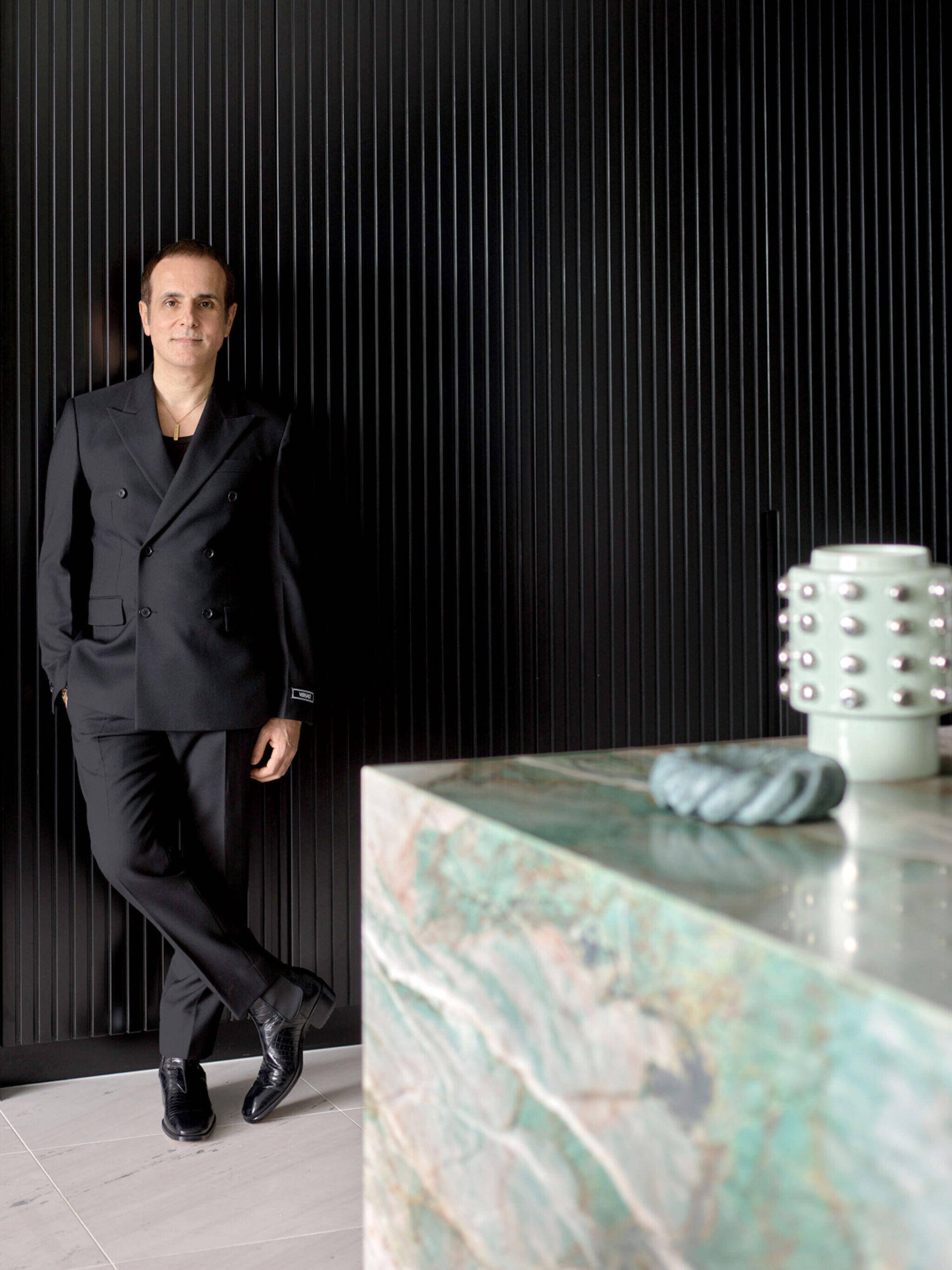 A person in a black suit stands against a black ribbed wall. In the foreground, a marble surface holds a mint green vase and a small blue object.