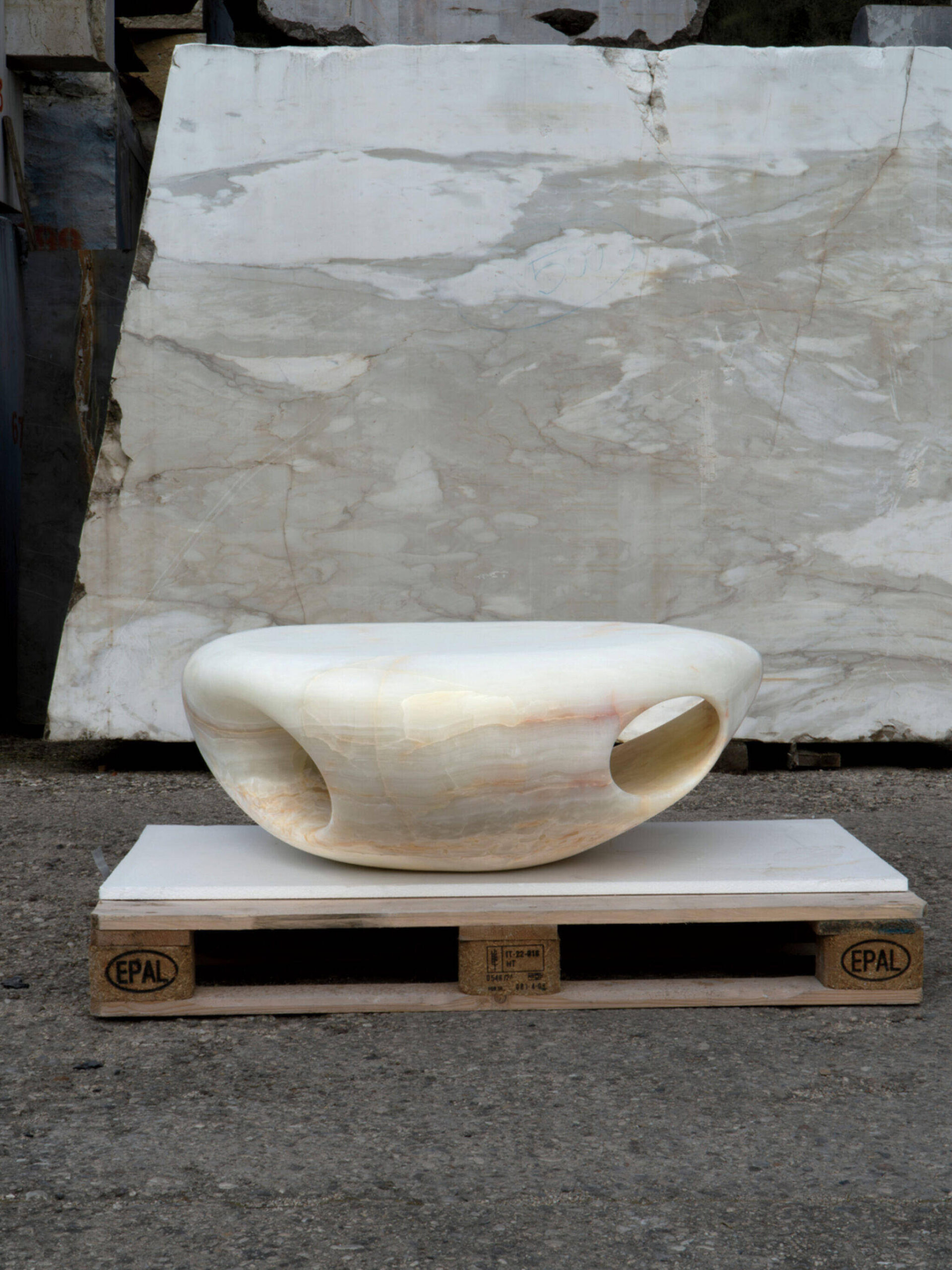 Smooth, white, oval stone sculpture with large rounded openings sits on a white platform, which is placed on two wooden pallets in an outdoor setting with marble slabs behind it.