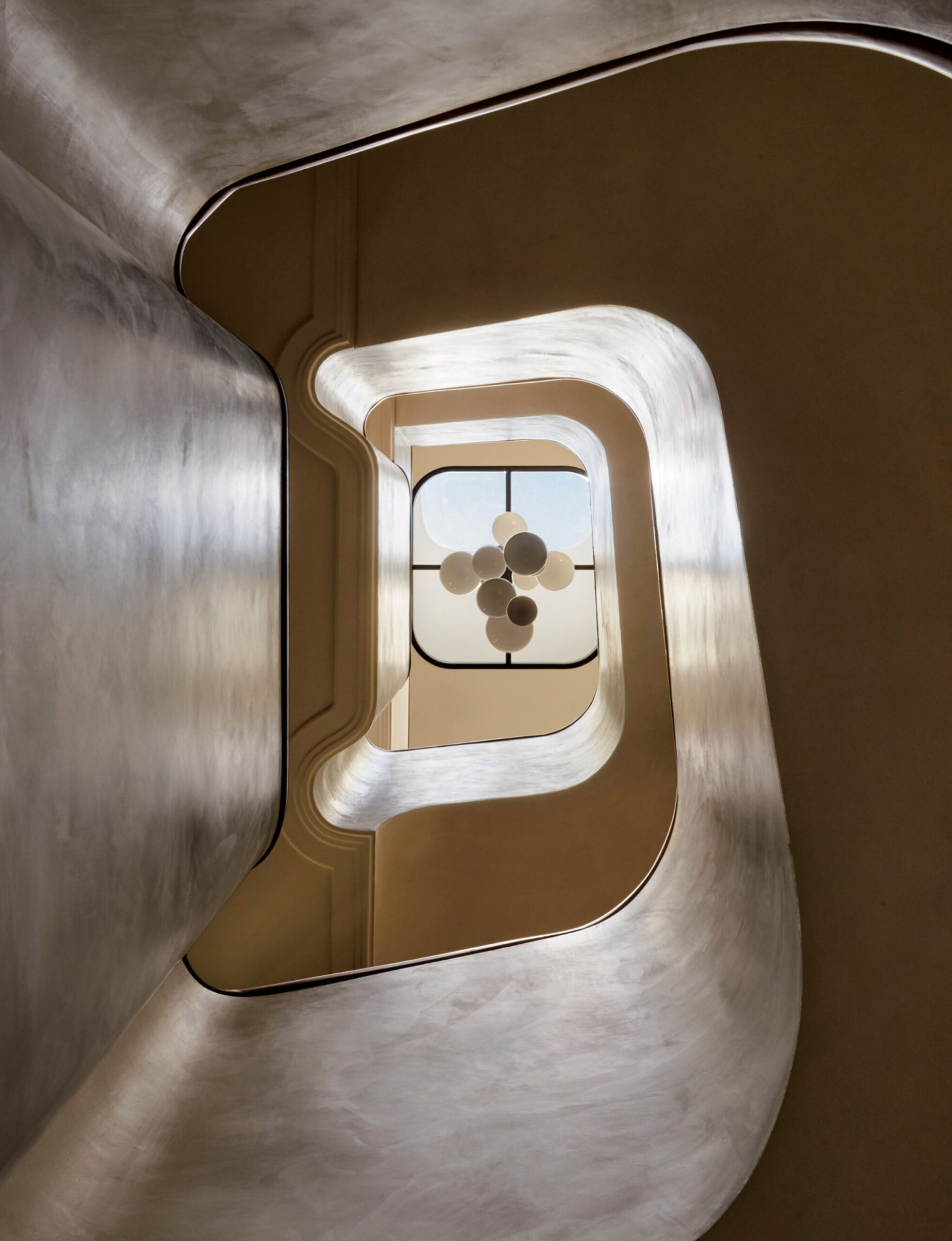 View looking up a modern spiral staircase with smooth, curved walls and a geometric chandelier hanging below a window at the top.