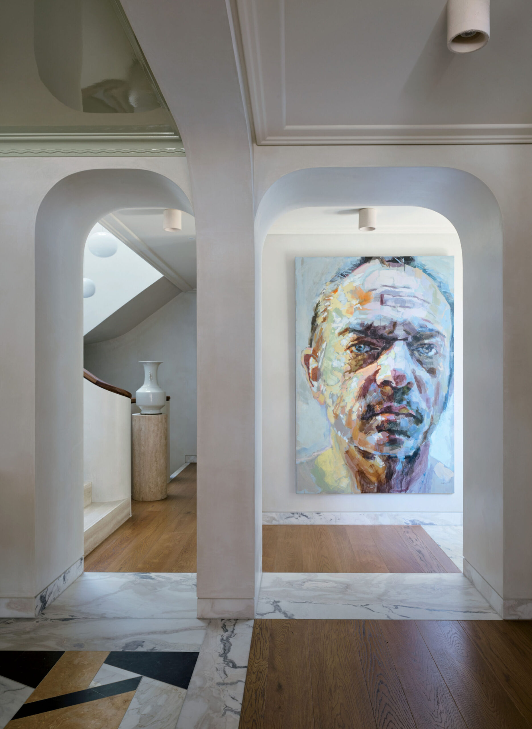 A hallway with marble and wood floors leads to a large, colorful portrait of a man’s face hanging on a wall; a white vase sits on a ledge beside a staircase.