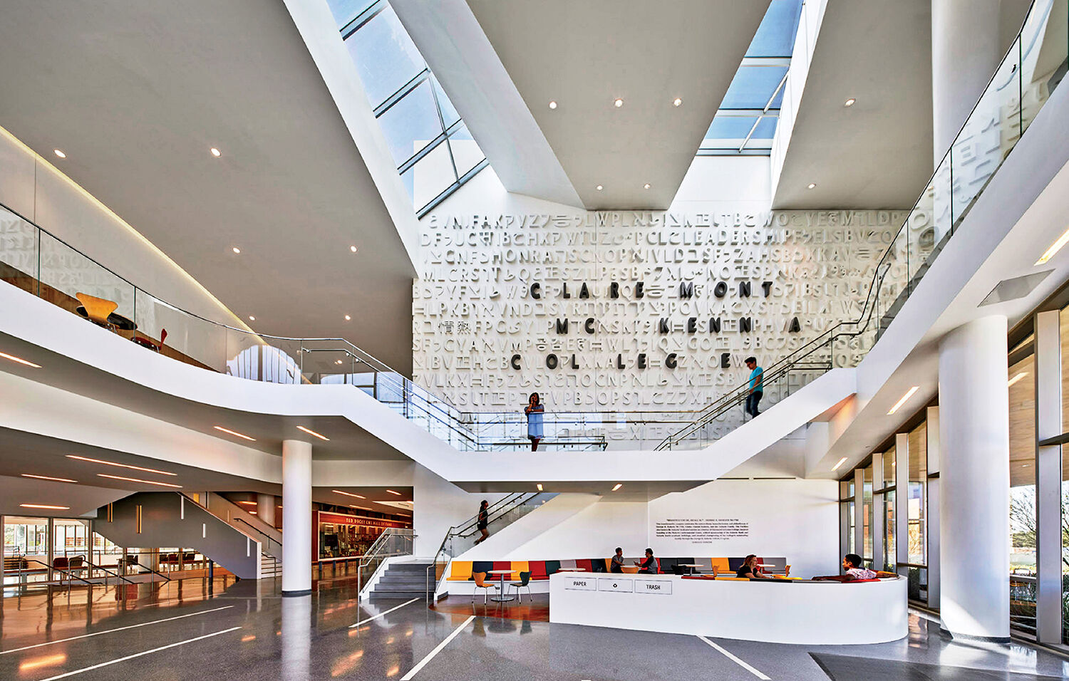 Modern building lobby with high ceilings, skylights, a lettered wall, white walls, a main desk, couches, and people walking on upper and lower levels.