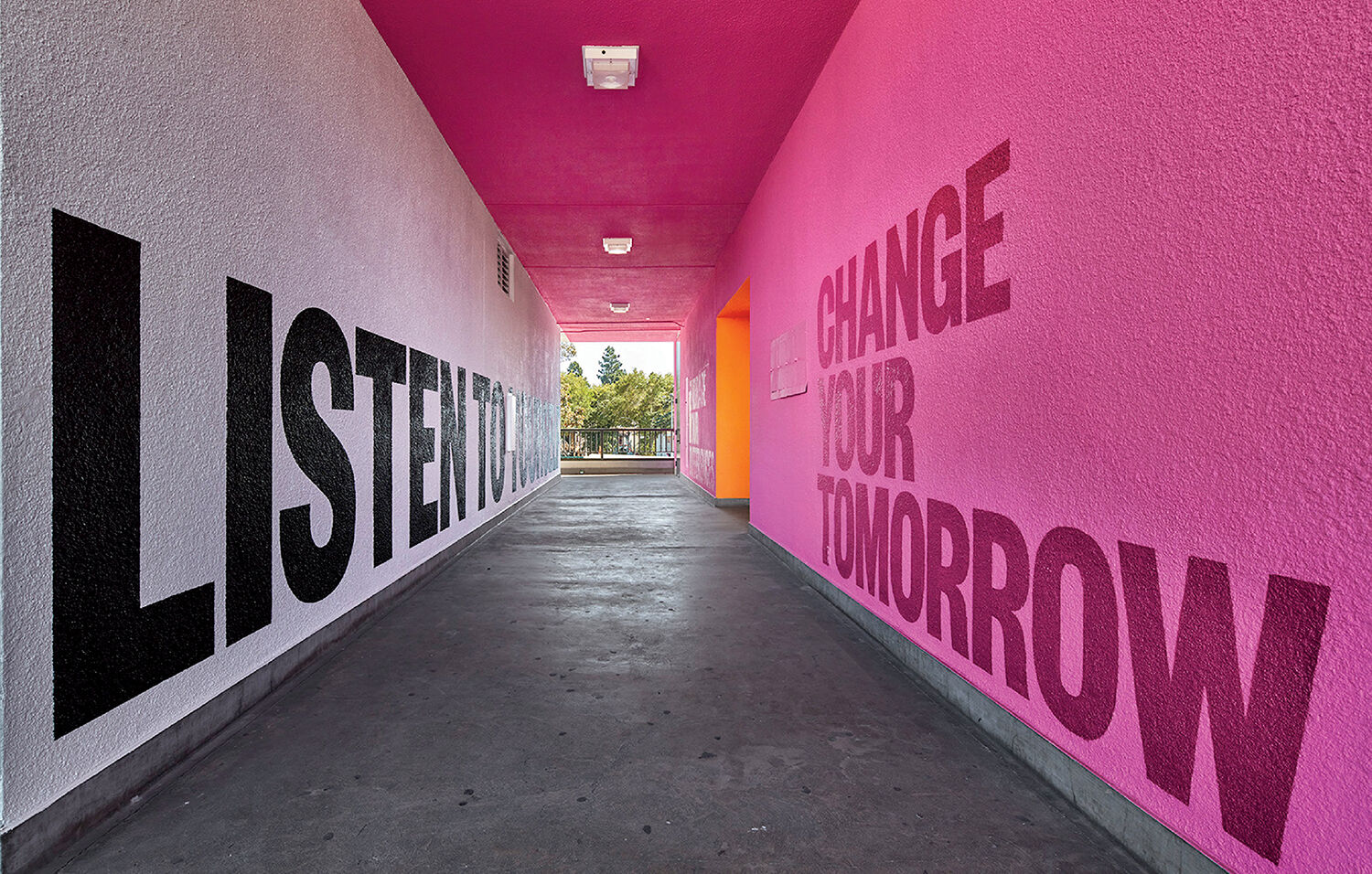 A corridor with pink walls features large black and pink text that reads, “LISTEN TO” on the left and “CHANGE YOUR TOMORROW” on the right. The ceiling is also painted bright pink.