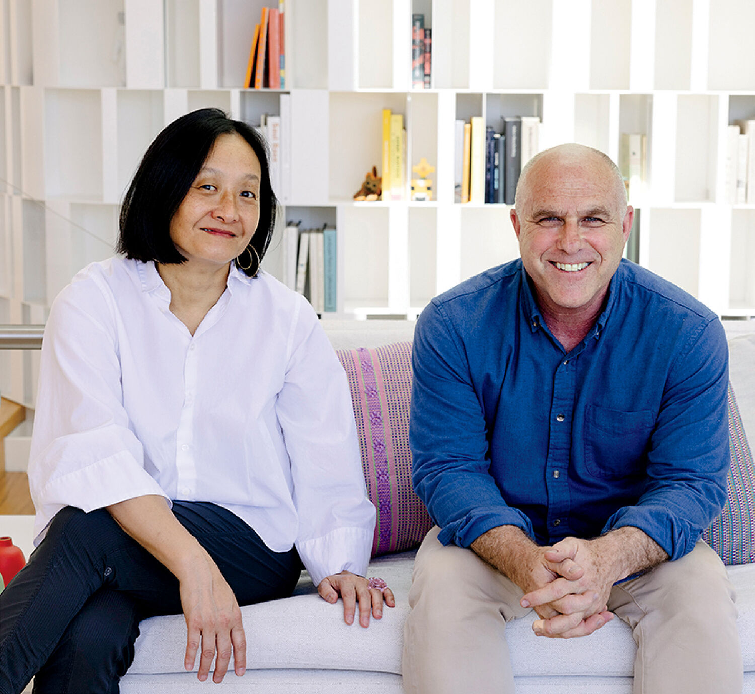 Two people sitting on a white couch in front of a bookshelf, both facing the camera and smiling.
