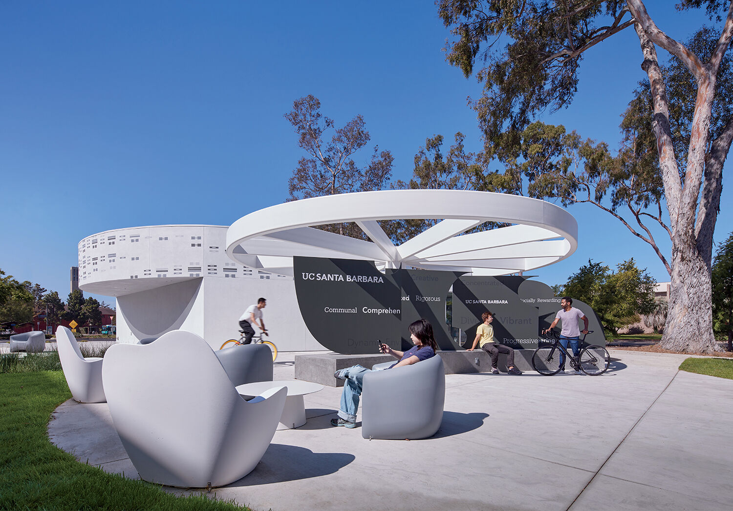 Several people sit, walk, and ride bikes near a modern outdoor seating area and a large UC Santa Barbara sign, with trees and campus buildings in the background.