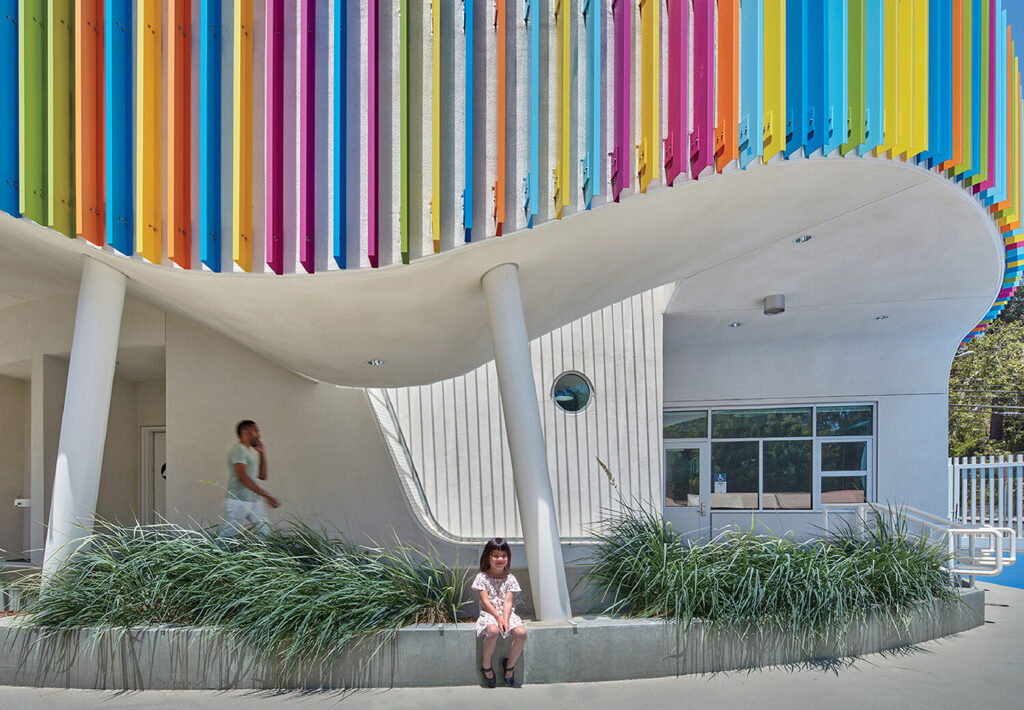 A young girl sits on a ledge in front of a modern white building with colorful vertical panels. An adult walks in the background.