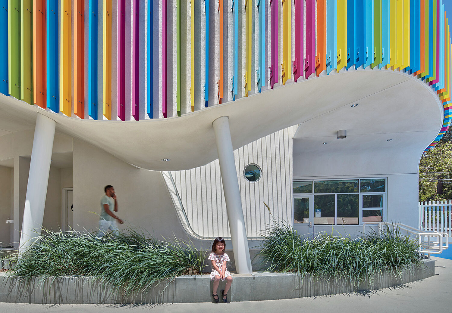 A young girl sits on a ledge in front of a modern white building with colorful vertical panels. An adult walks in the background.