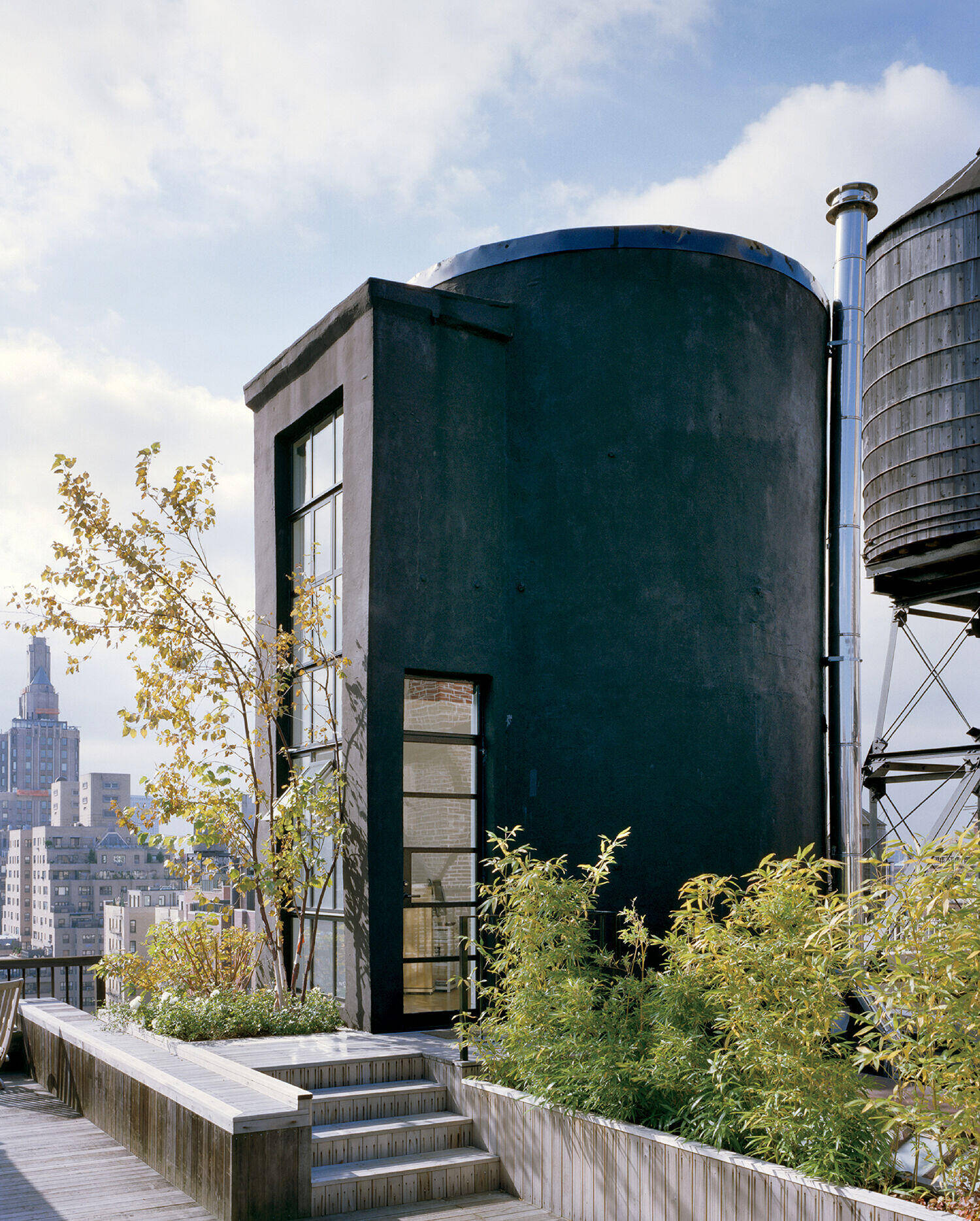 A modern black cylindrical structure with large windows sits on a rooftop garden with city buildings and a water tower in the background.