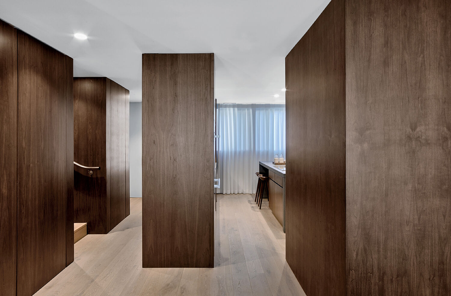 Modern interior with large vertical wooden panels, light wood flooring, and soft natural light from a window with sheer curtains; part of a kitchen area is visible in the background.