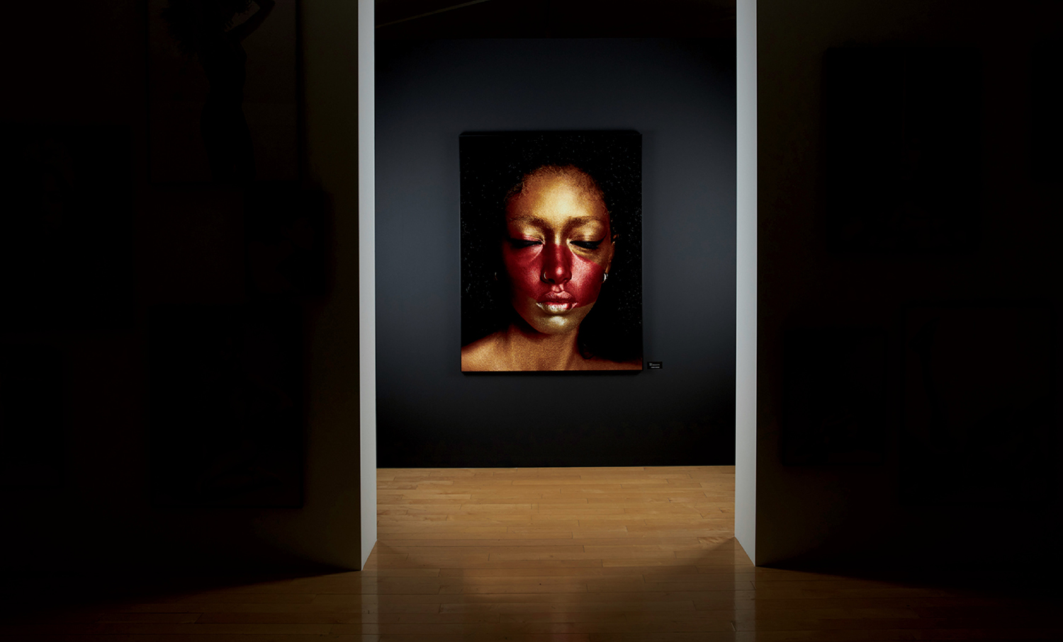A portrait of a person with dramatic red and gold face paint is displayed on a dark wall in a dimly lit gallery space.