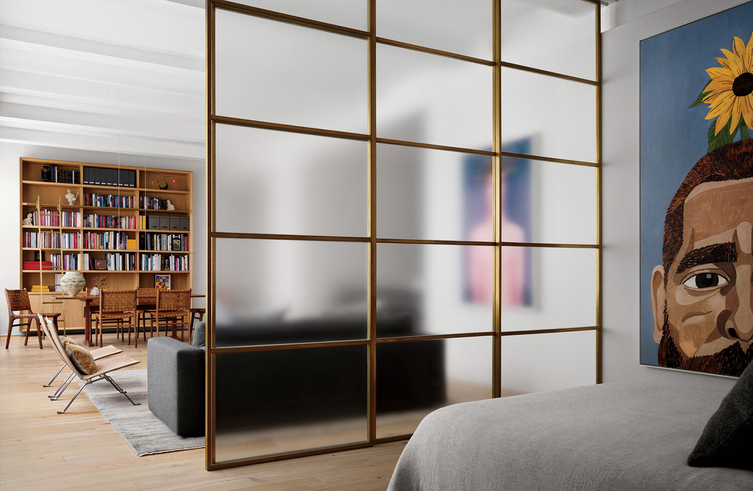 Modern open-plan living space with wooden floors, a frosted glass room divider, bookshelves, dining table, and artwork featuring a man’s face with a sunflower on his head.