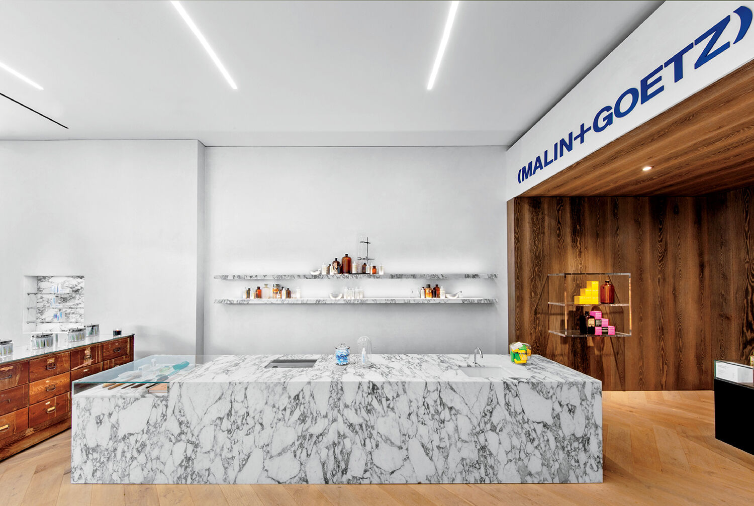 Modern retail store interior featuring a marble display counter, minimalist shelves with products, wood paneling, and a MALIN+GOETZ sign.