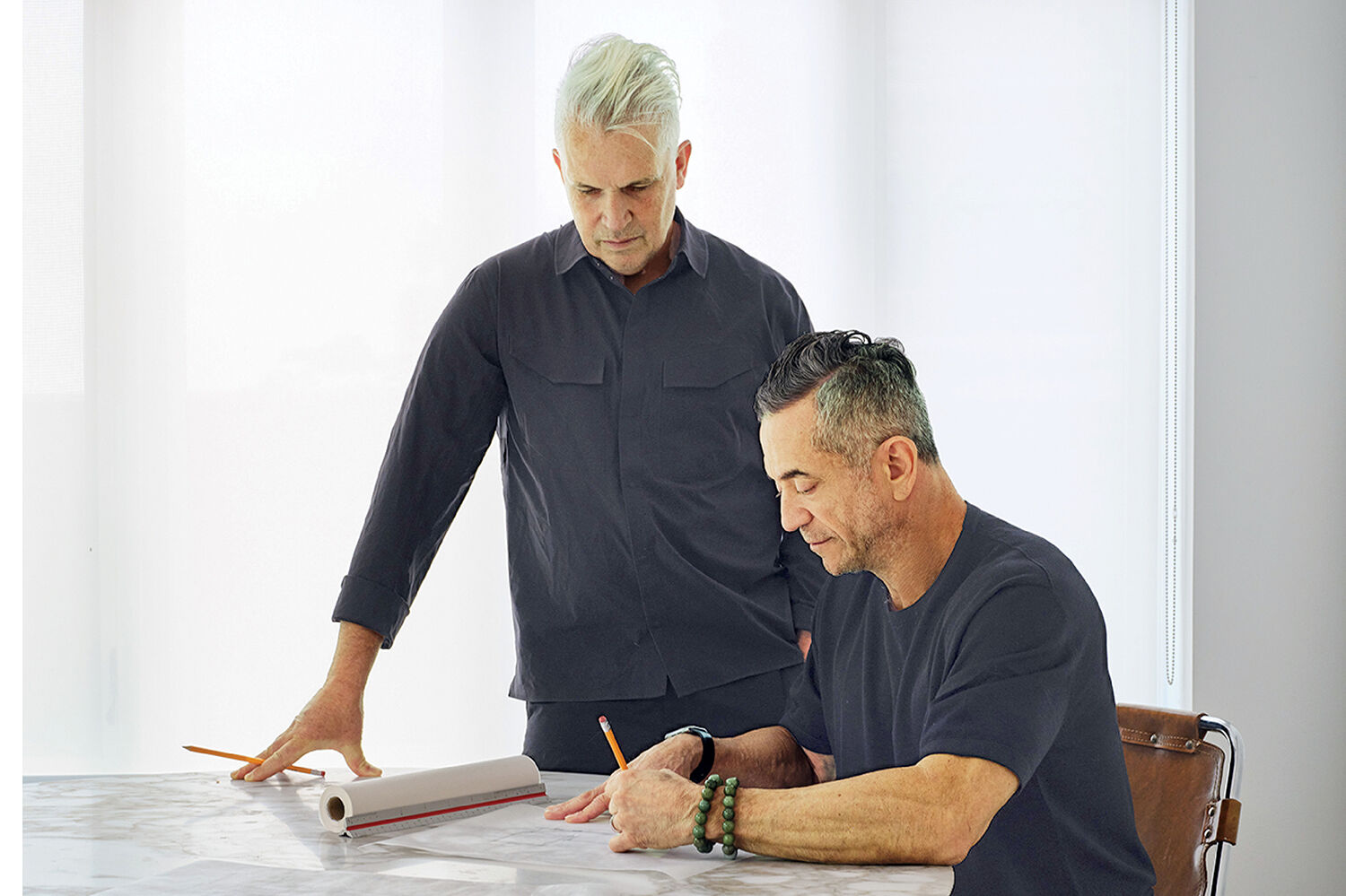 Two men at a table look over documents; one is standing with a pencil and the other is sitting and writing.