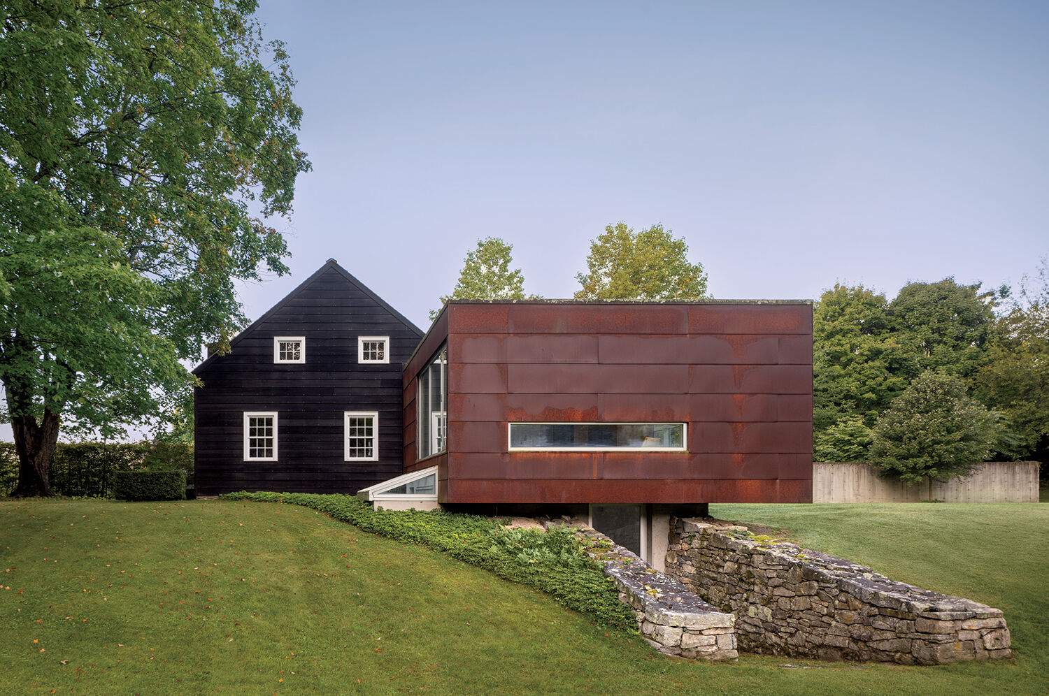 A modern extension with rust-colored metal siding projects from a traditional dark house, set on a grassy lawn with trees and a stone retaining wall.