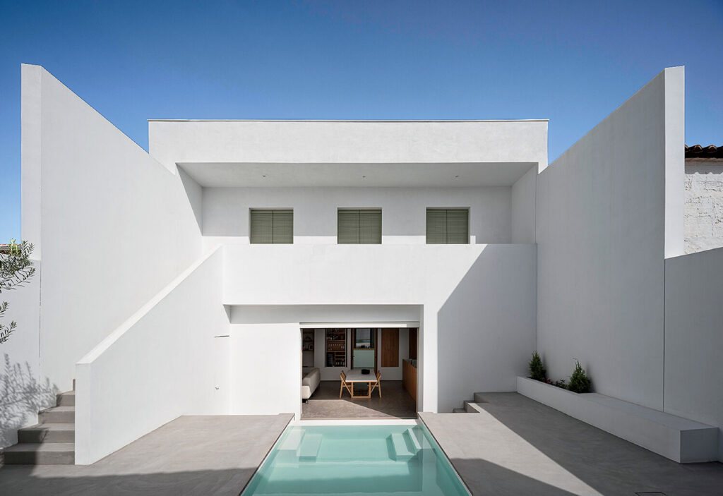 Modern white minimalist house with flat roof, rectangular windows, and a central pool in the courtyard; doors open to a furnished interior, with stairs and planters on either side.