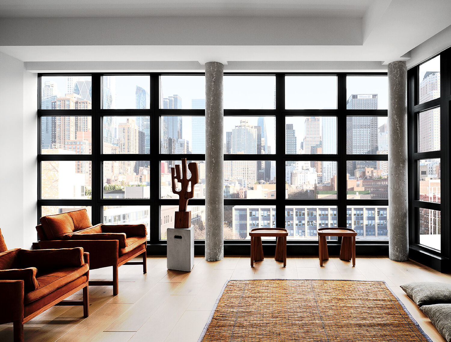 Modern living room with large grid windows, city skyline view, two armchairs, two wooden stools, a woven rug, and a cactus sculpture on a concrete pedestal.