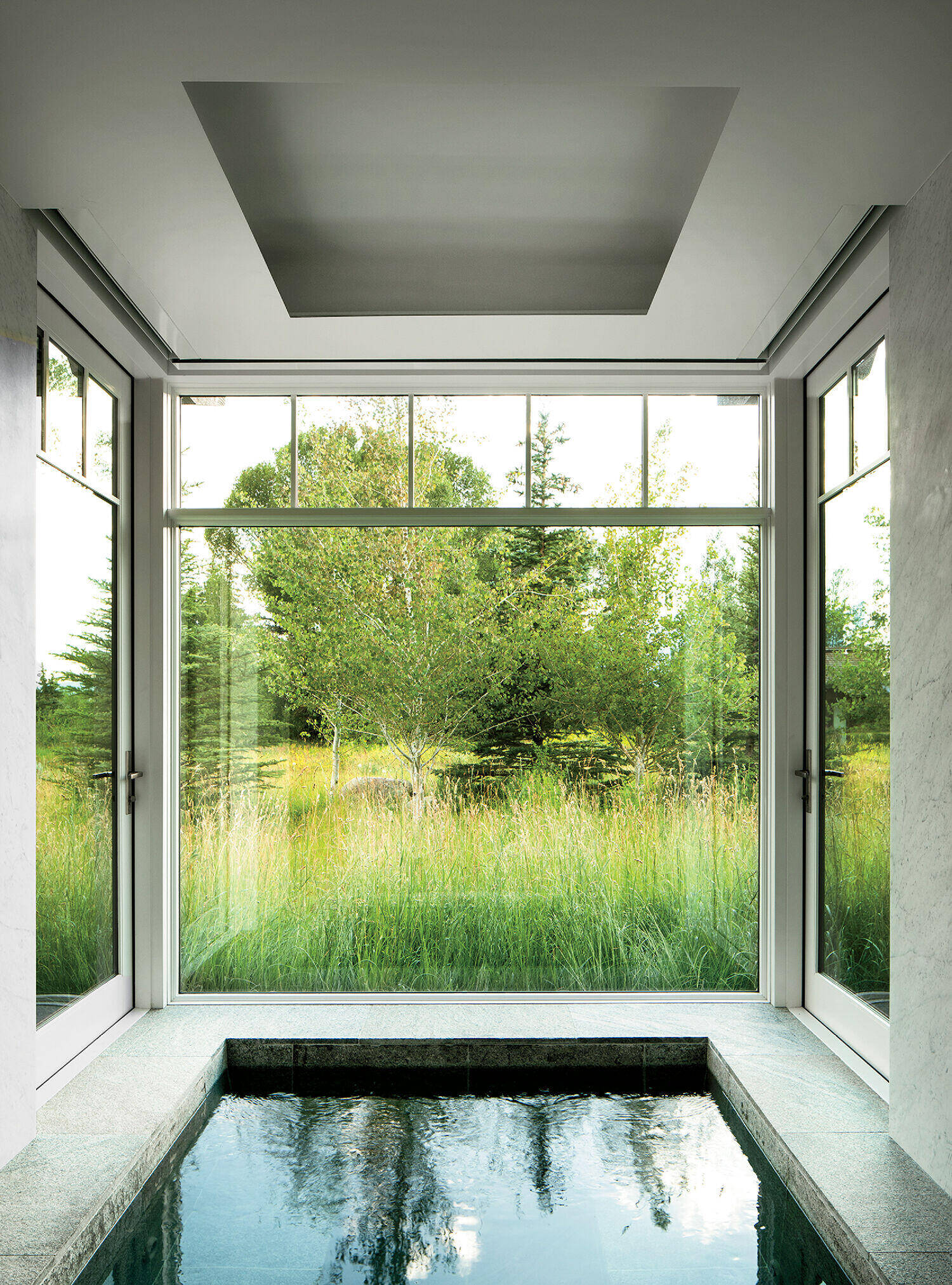 Indoor pool with marble edging, surrounded by large windows, overlooks a grassy landscape with trees in the background. Natural light fills the room.
