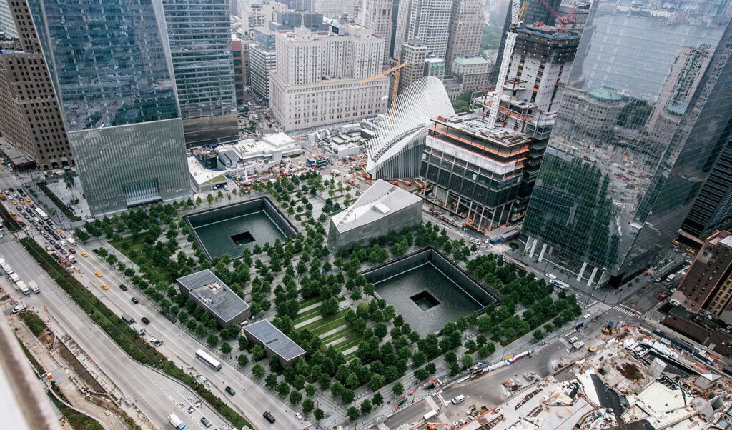 Aerial view of the 9/11 Memorial in New York City, showing two large square reflecting pools surrounded by trees and nearby skyscrapers.