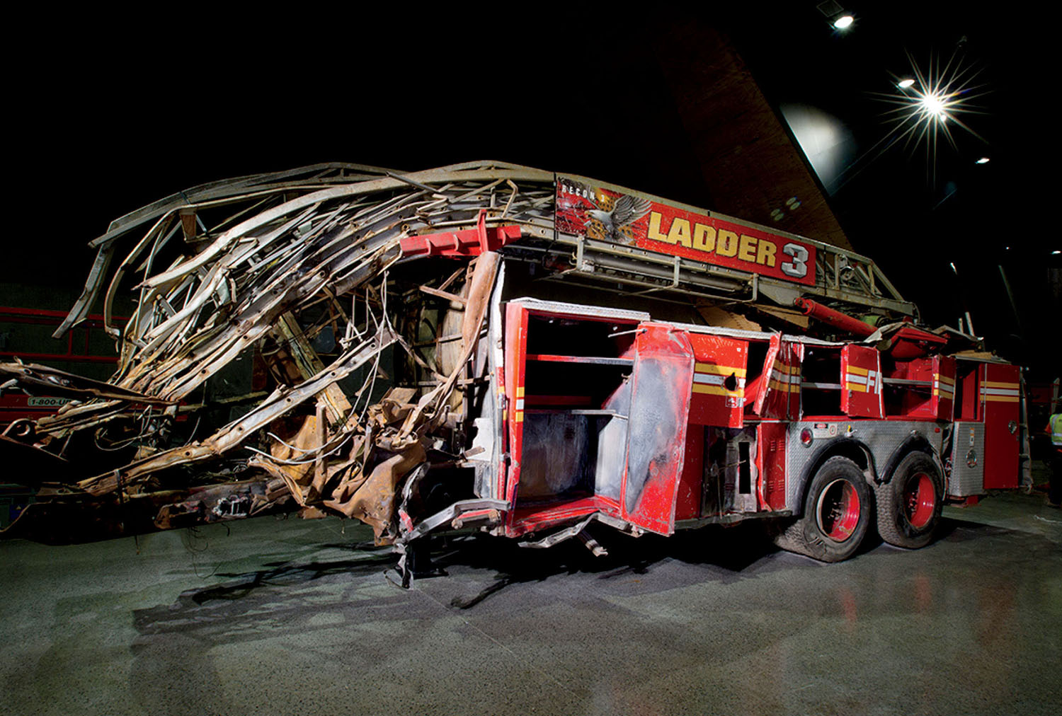 A heavily damaged fire truck labeled “Ladder 3” is displayed indoors under dim lighting, with twisted metal and open compartments visible.