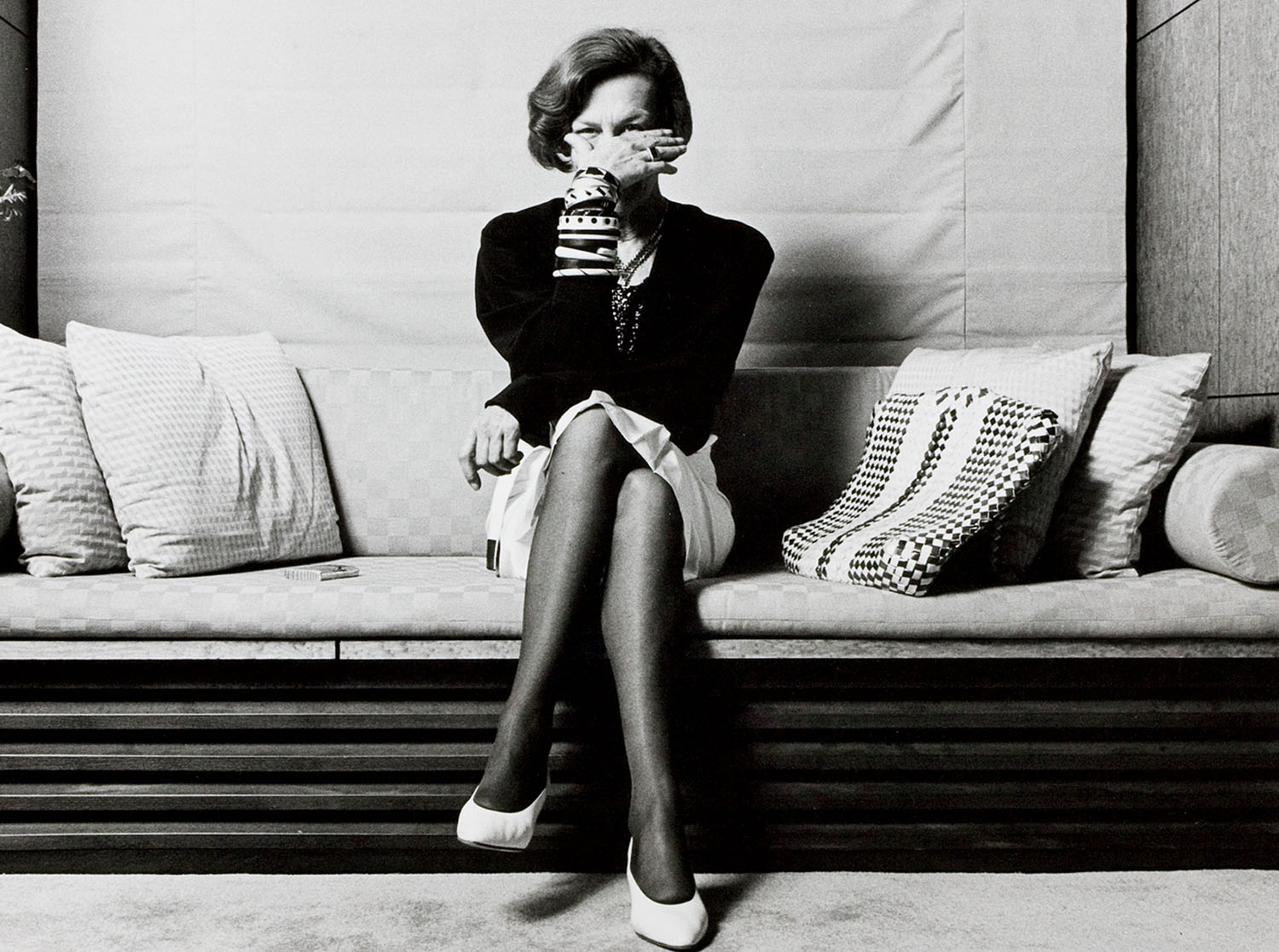 A woman sits cross-legged on a couch, holding a cup to her face, surrounded by patterned pillows in a modern room.