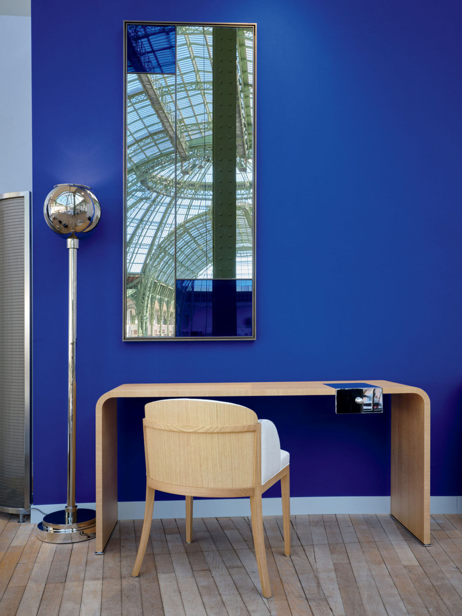 Minimalist workspace with a light wood desk, modern chair, floor lamp, and tall mirror against a vivid blue wall with wooden flooring.