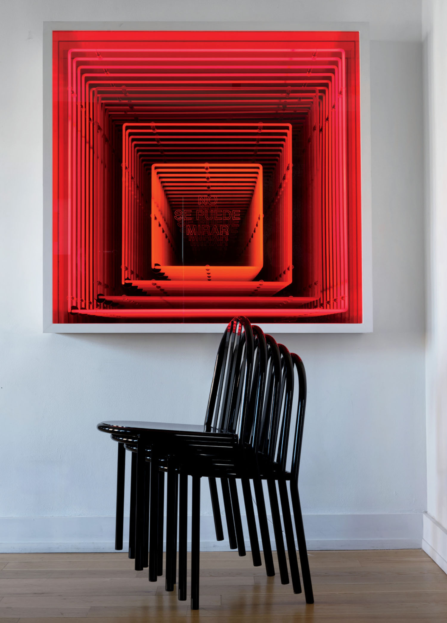 Four black chairs are stacked together in front of a red, geometric, tunnel-like wall artwork with text at the center, in a minimalist room with white walls and floor.