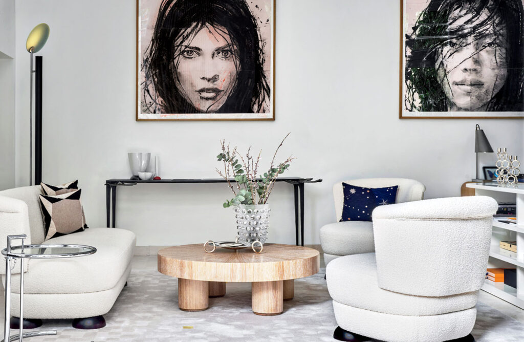 Modern living room with two white chairs, a round wooden coffee table, wall art featuring two faces, and minimalist decor on a console table against a white wall.