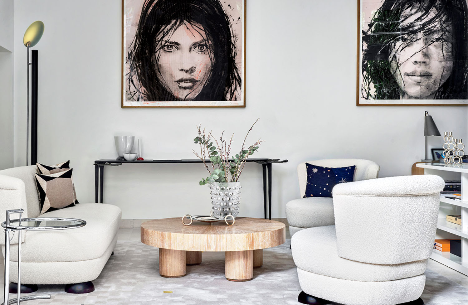 Modern living room with two white chairs, a round wooden coffee table, wall art featuring two faces, and minimalist decor on a console table against a white wall.