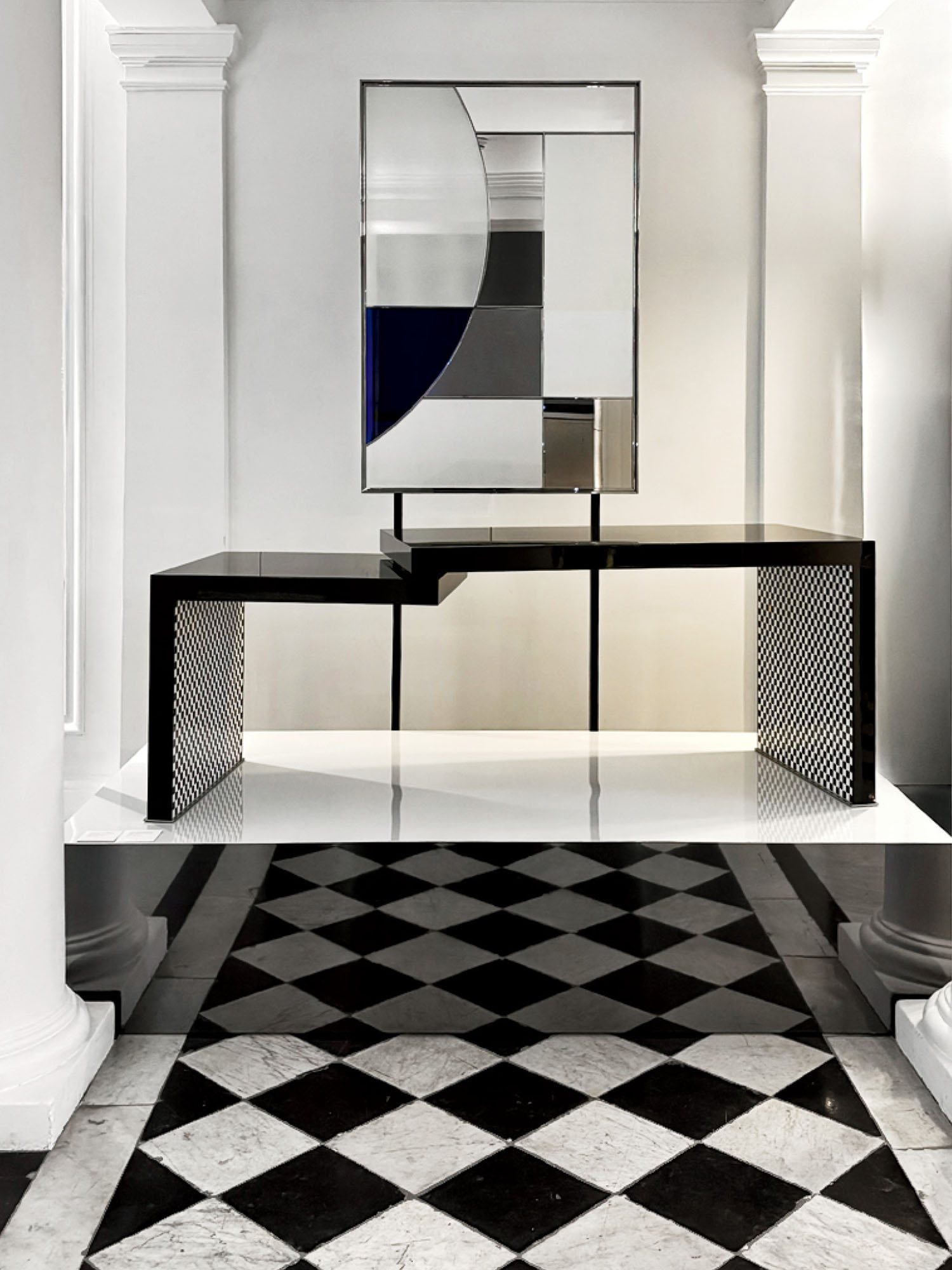 A modern black table with geometric legs stands on a black and white checkered floor, with a rectangular abstract mirror placed above it between two white columns.