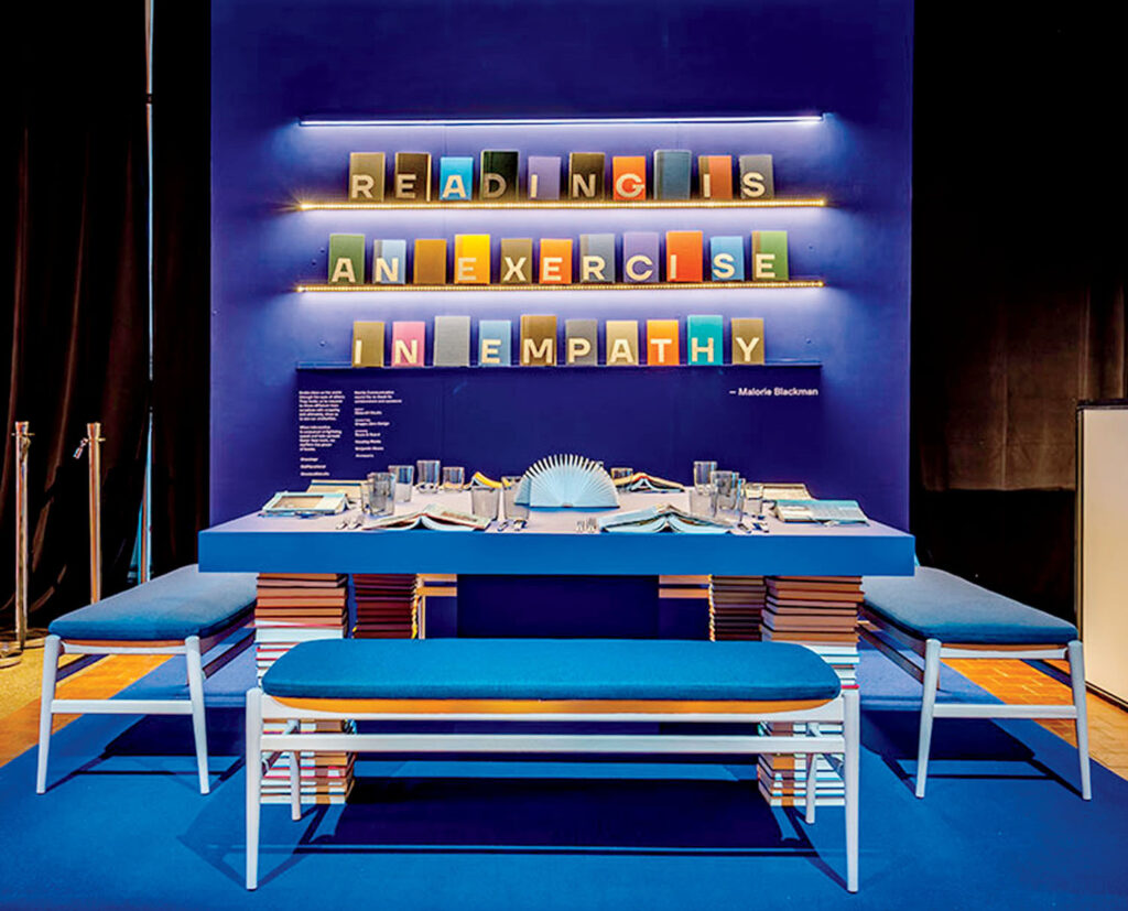 A blue table set for dining, with books stacked as table legs and wall shelves spelling, “Reading is an exercise in empathy,” against a dark blue backdrop.