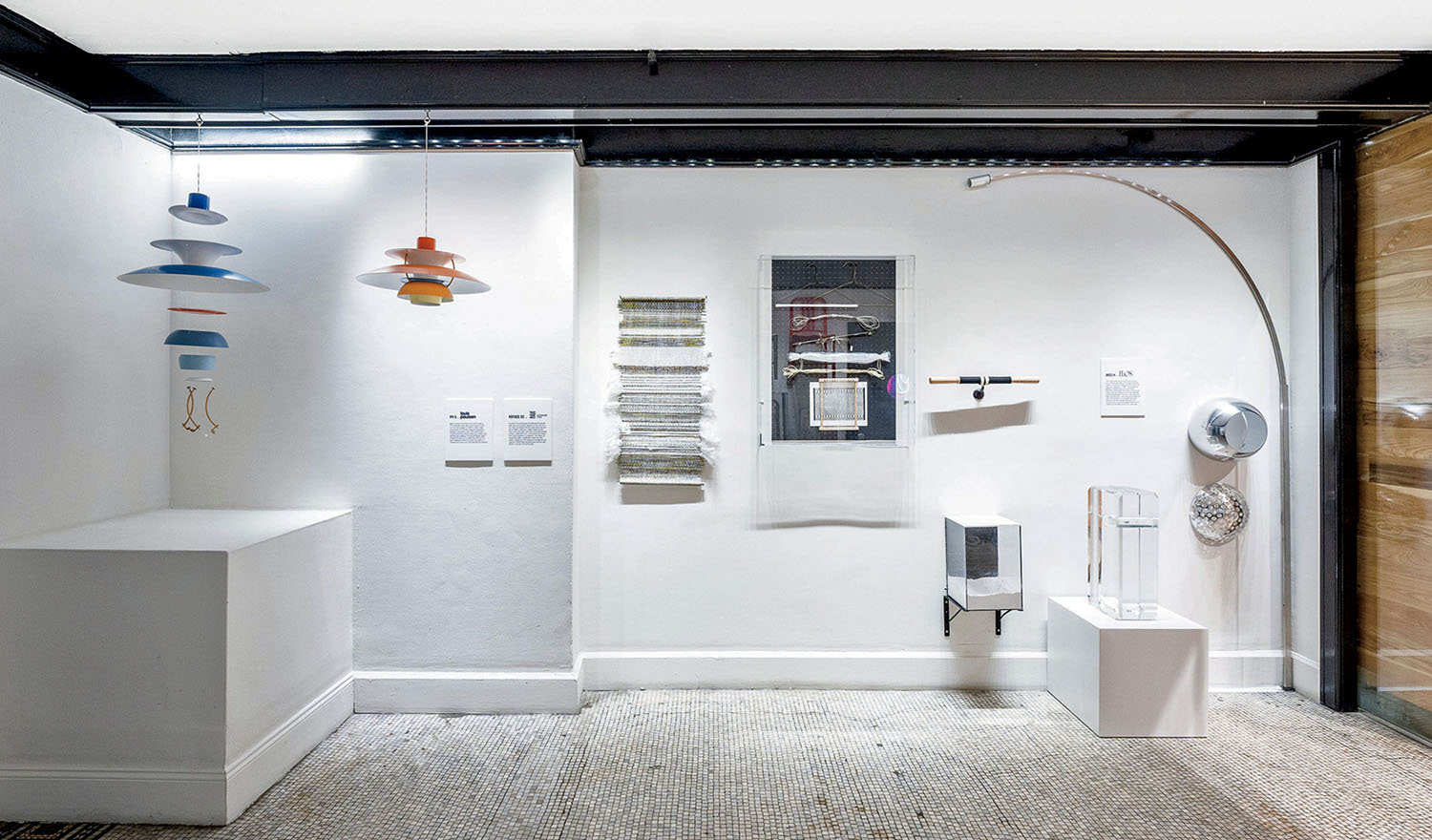 Exhibit display with modern hanging lamps, woven textile panels, sculptural objects on pedestals, and descriptive wall text in a well-lit gallery space.