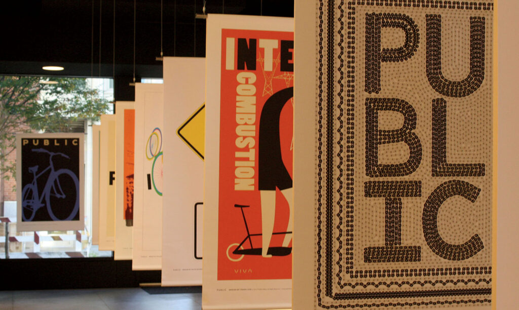 A row of colorful poster prints hangs in a modern gallery, with the foreground poster displaying the word “PUBLIC” in bold letters.