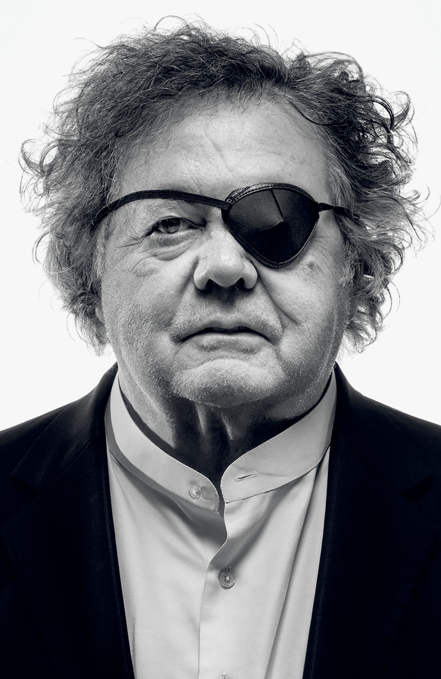 Older man with curly hair wearing a dark eyepatch, suit jacket, and buttoned shirt, looking directly at the camera with a neutral expression against a plain background.
