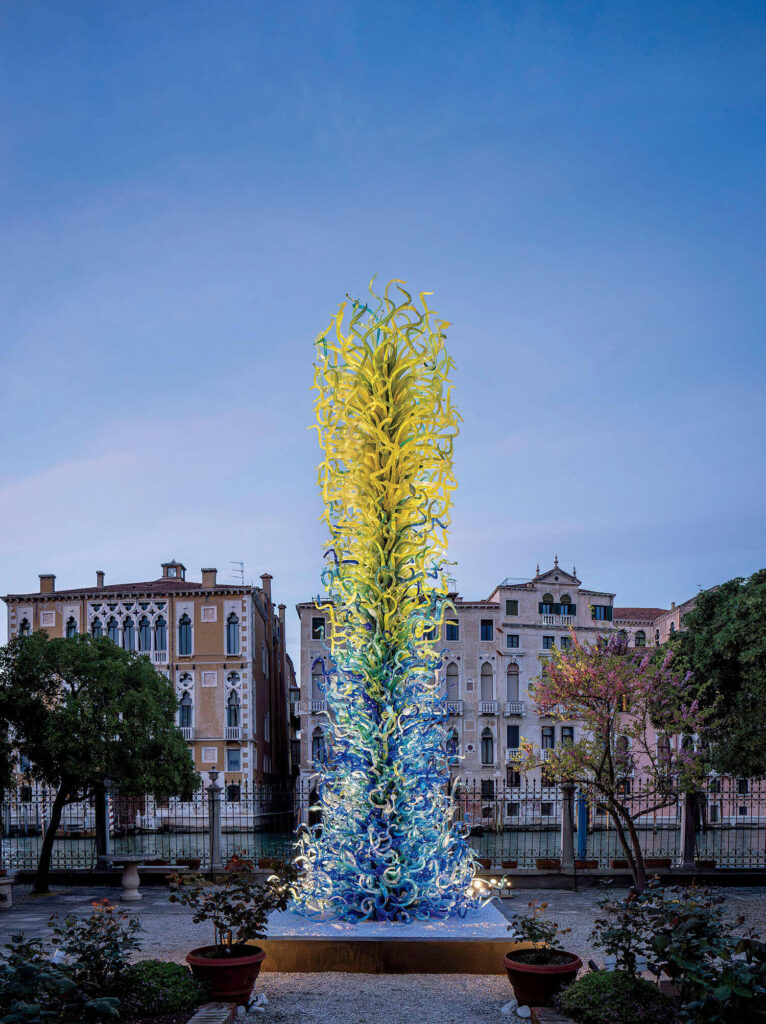 Tall glass sculpture with yellow and blue twisting shapes stands in a garden, surrounded by trees and historic buildings under a clear sky.