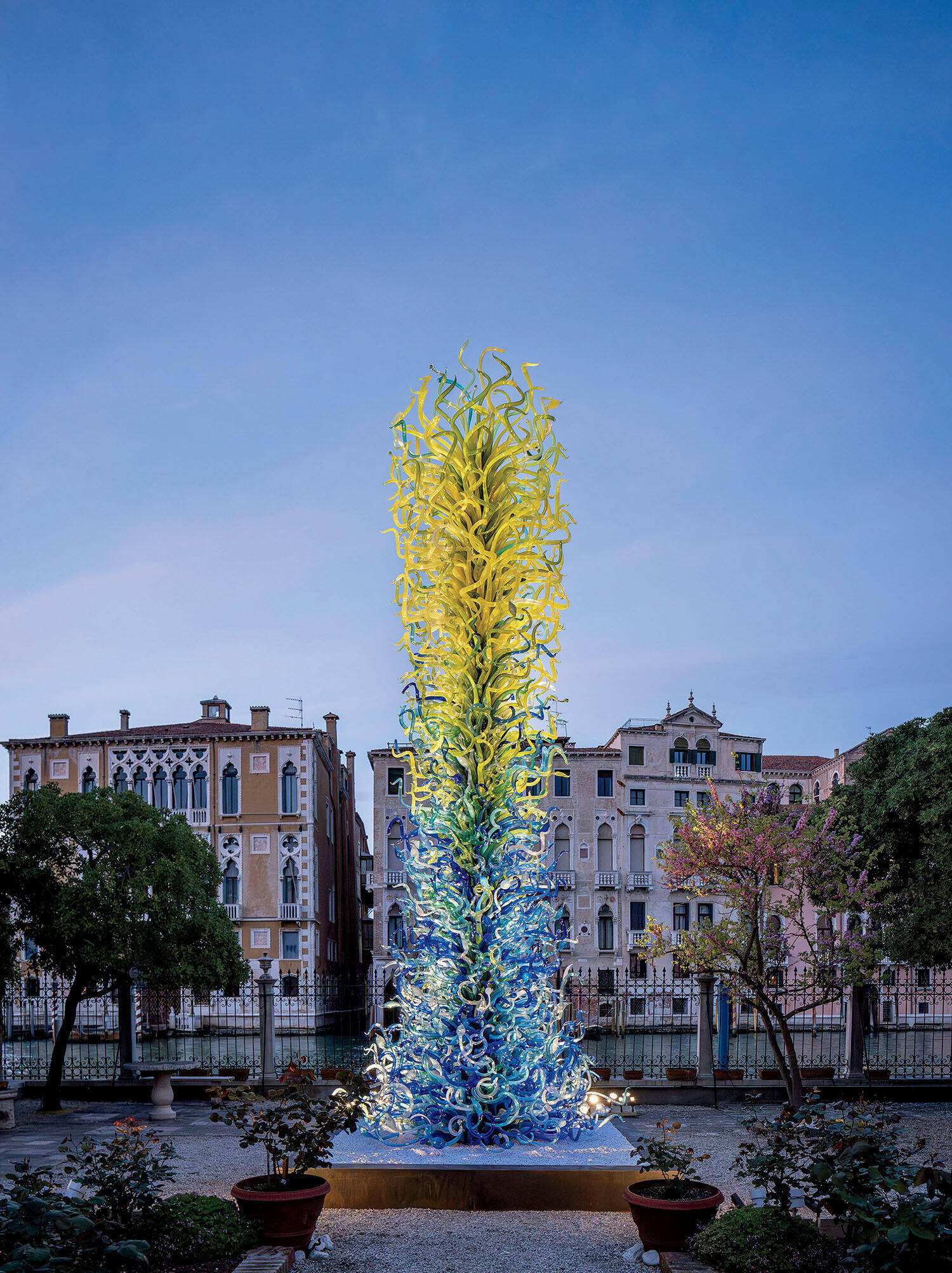 Tall glass sculpture with yellow and blue twisting shapes stands in a garden, surrounded by trees and historic buildings under a clear sky.