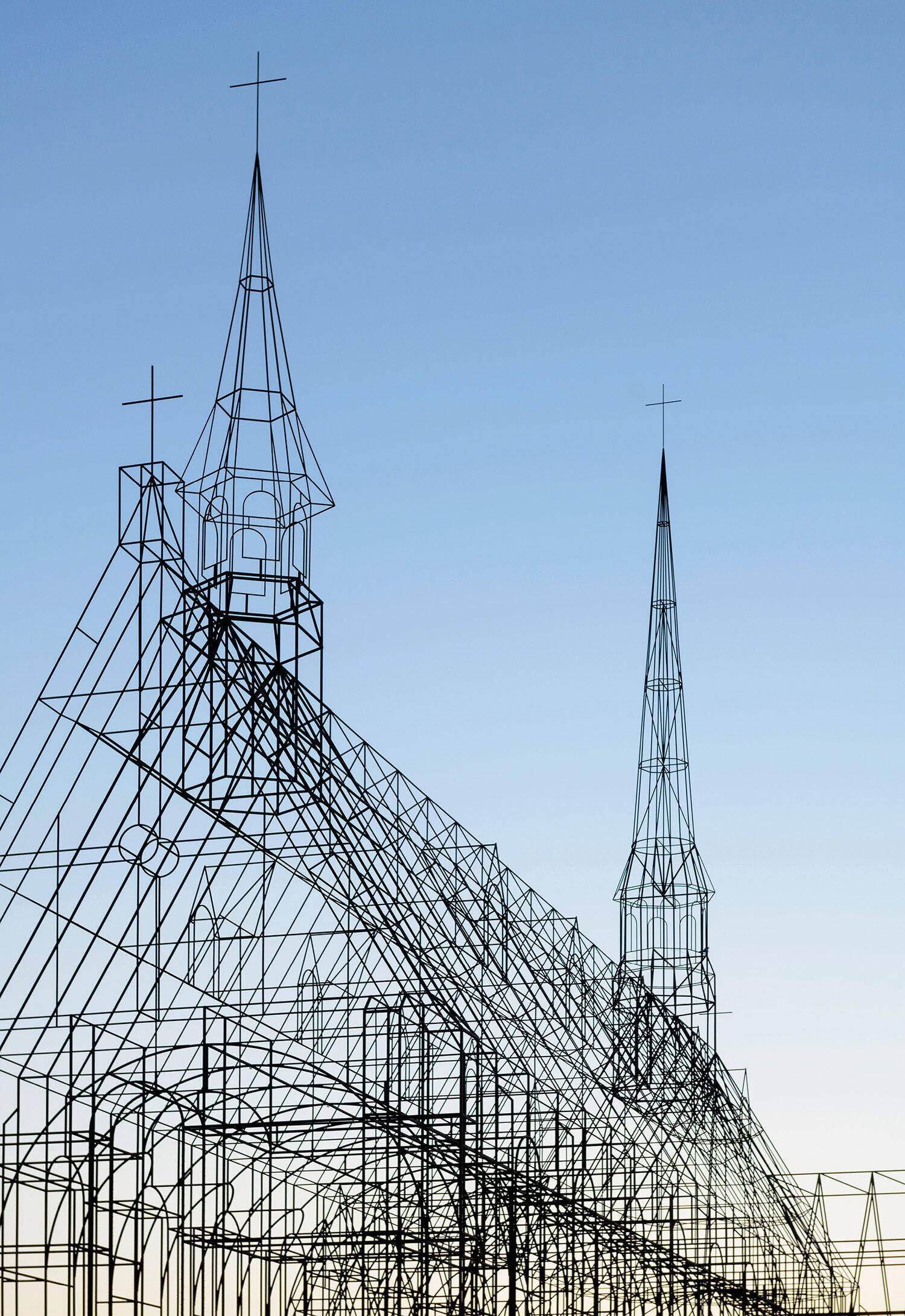 Wireframe structure resembling a church with pointed steeples and crosses, set against a clear blue sky at sunset.