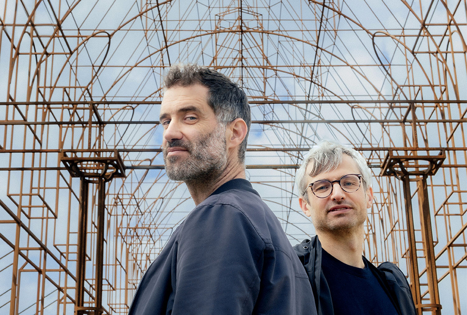 Two men stand in front of a large metal geometric structure with intersecting lines, looking at the camera under a partly cloudy sky.