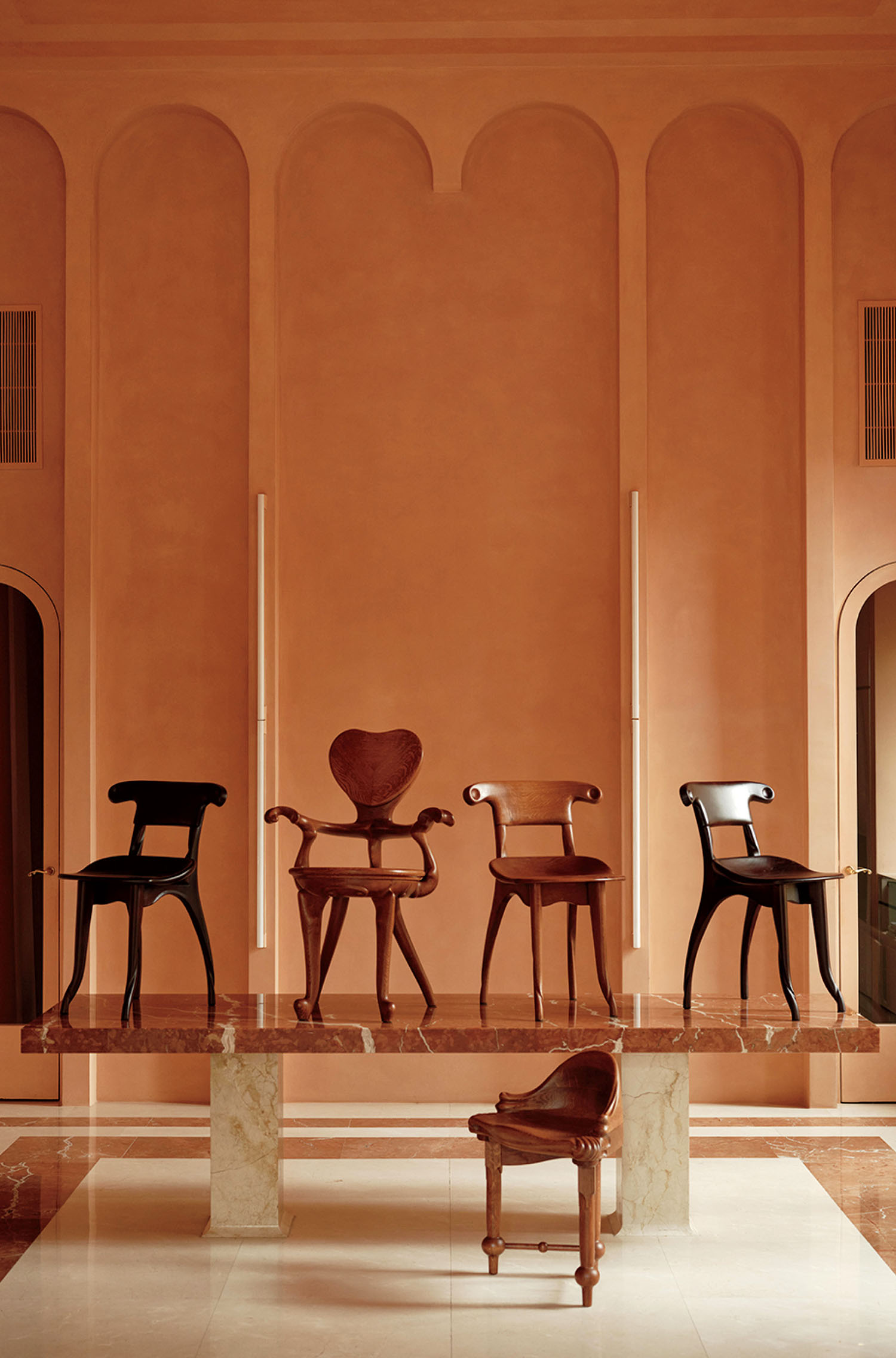 Five uniquely designed wooden chairs are displayed on a marble platform in an elegant, peach-toned room with arched wall details.