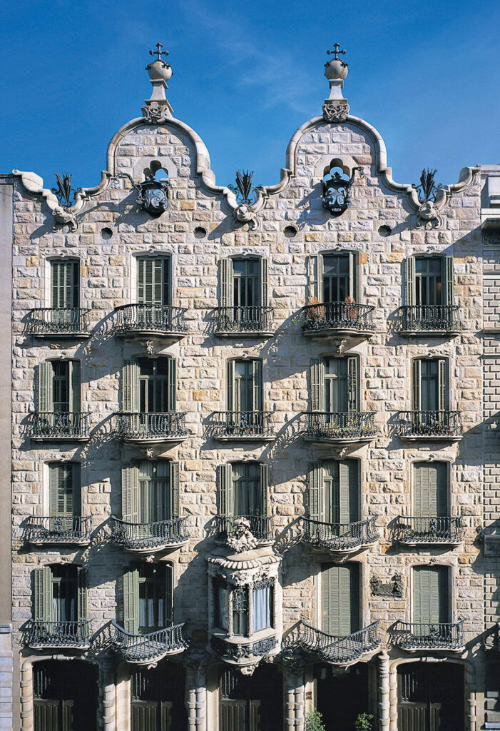 Ornate stone building façade with curved gables, iron balconies, and detailed architectural features, photographed under a clear blue sky.